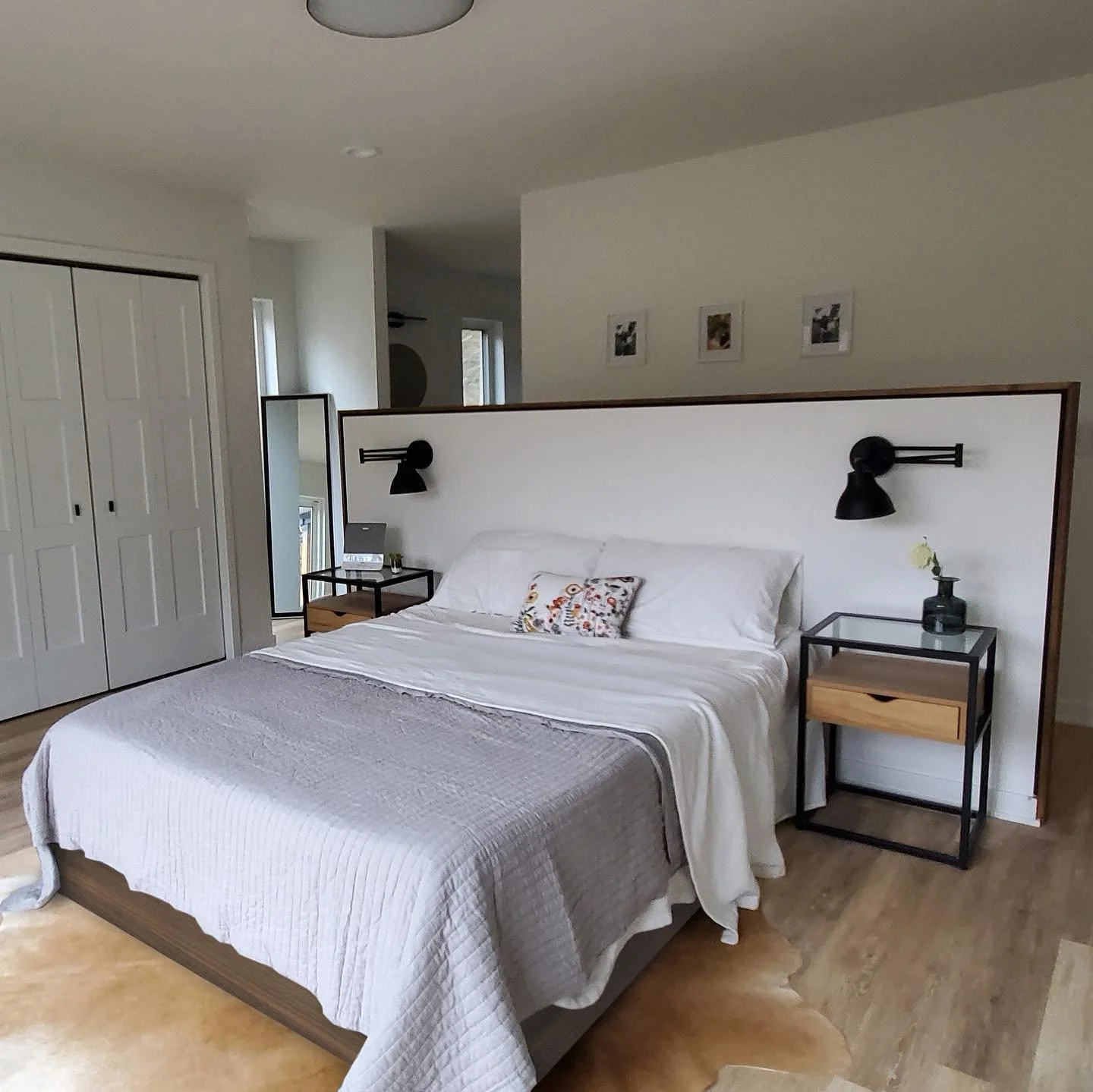 A bedroom with a bed, white bedding, and two black wall-mounted lamps above black bedside tables with natural wood drawers. There is a full-length mirror, a small table with a framed photo or art, and a mirror on a stand. The room has light-colored wooden flooring and a white wall with three framed photographs.