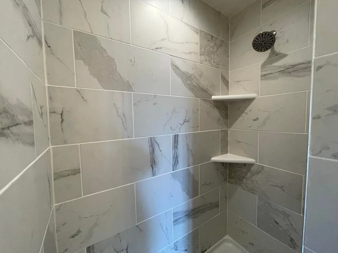Photo of a shower with marble-style tile walls and two corner shelves. A round shower head is visible in the upper right corner.