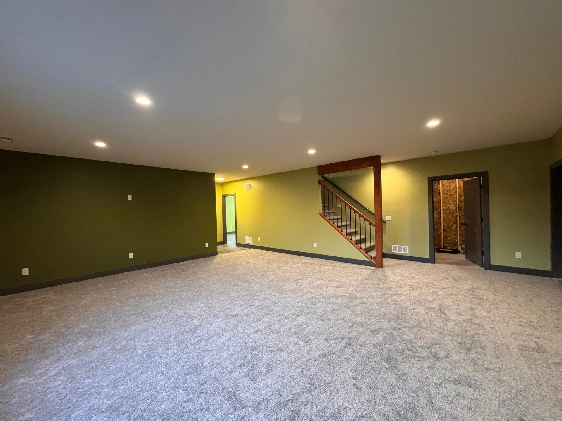 A family room with green walls, beige carpet, and recessed lighting. There's a staircase with wooden railing and black metal balusters, and doorways leading to other rooms.
