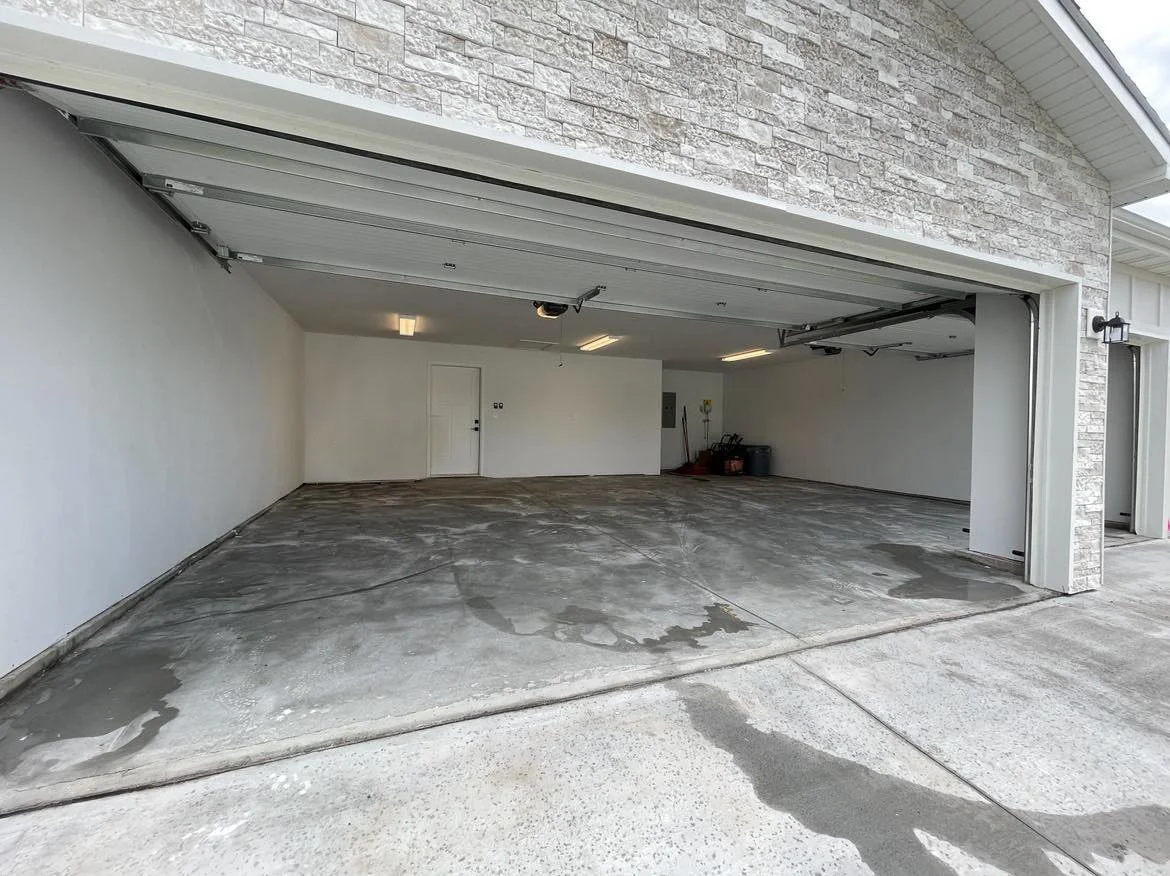 A two-car garage with a concrete floor, white walls, and a closed garage door. Some tools and supplies are stored in the back corner.