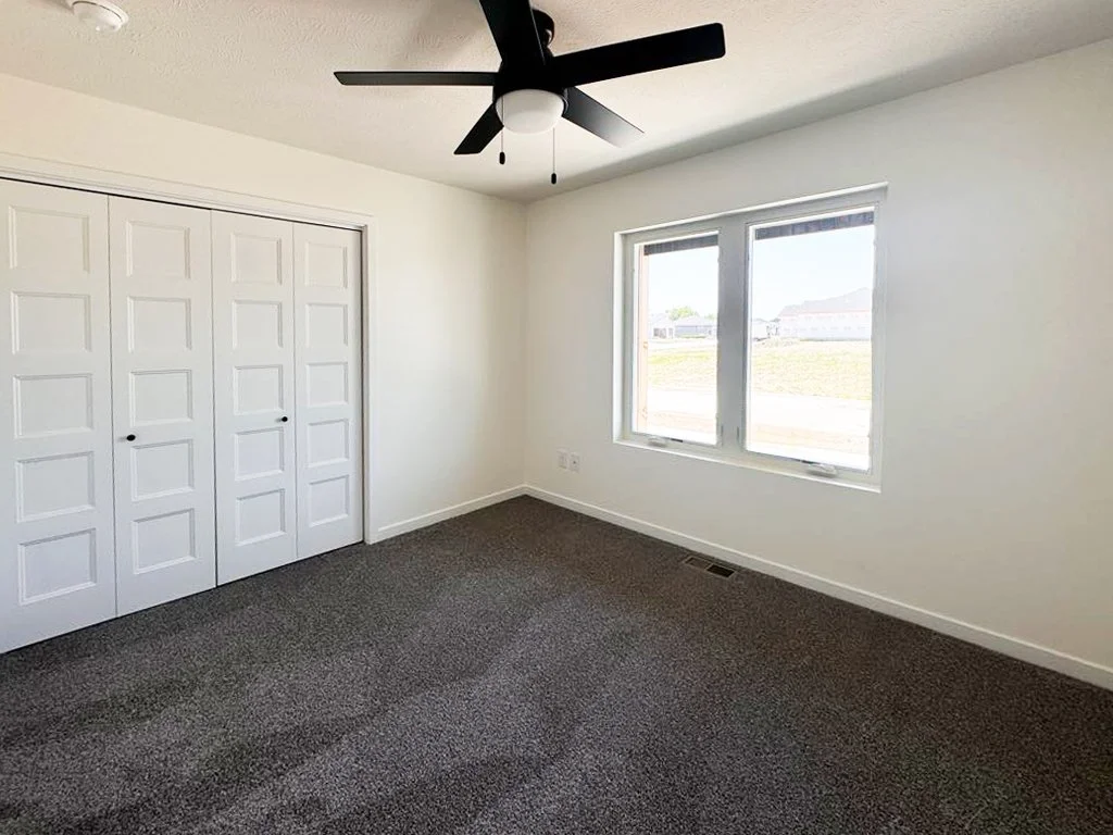 A bedroom with white walls, a window showing outside, a ceiling fan with a light, and a closet with white bi-fold doors.