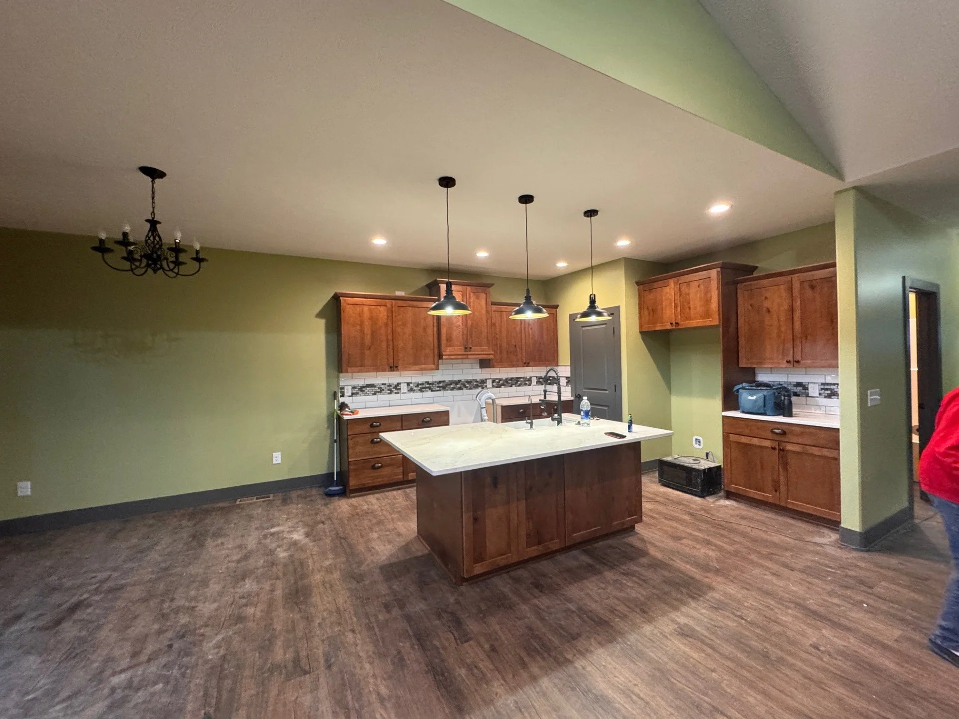 A kitchen with wooden cabinets, a kitchen island, pendant lights, and a chandelier, with a person wearing a red jacket partially visible on the right.