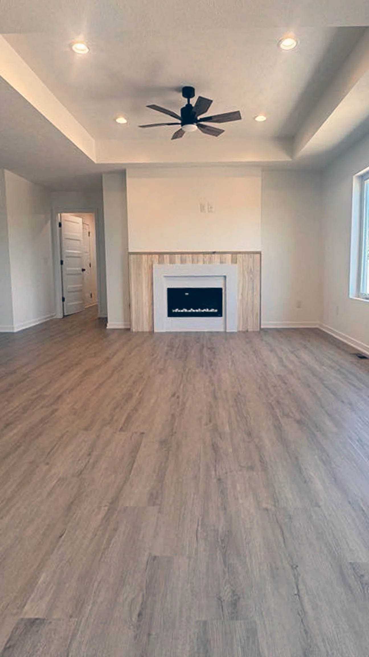 A living room with hardwood floors, white walls, a ceiling fan, recessed lighting, a fireplace with a wooden surround, and a window to the right.