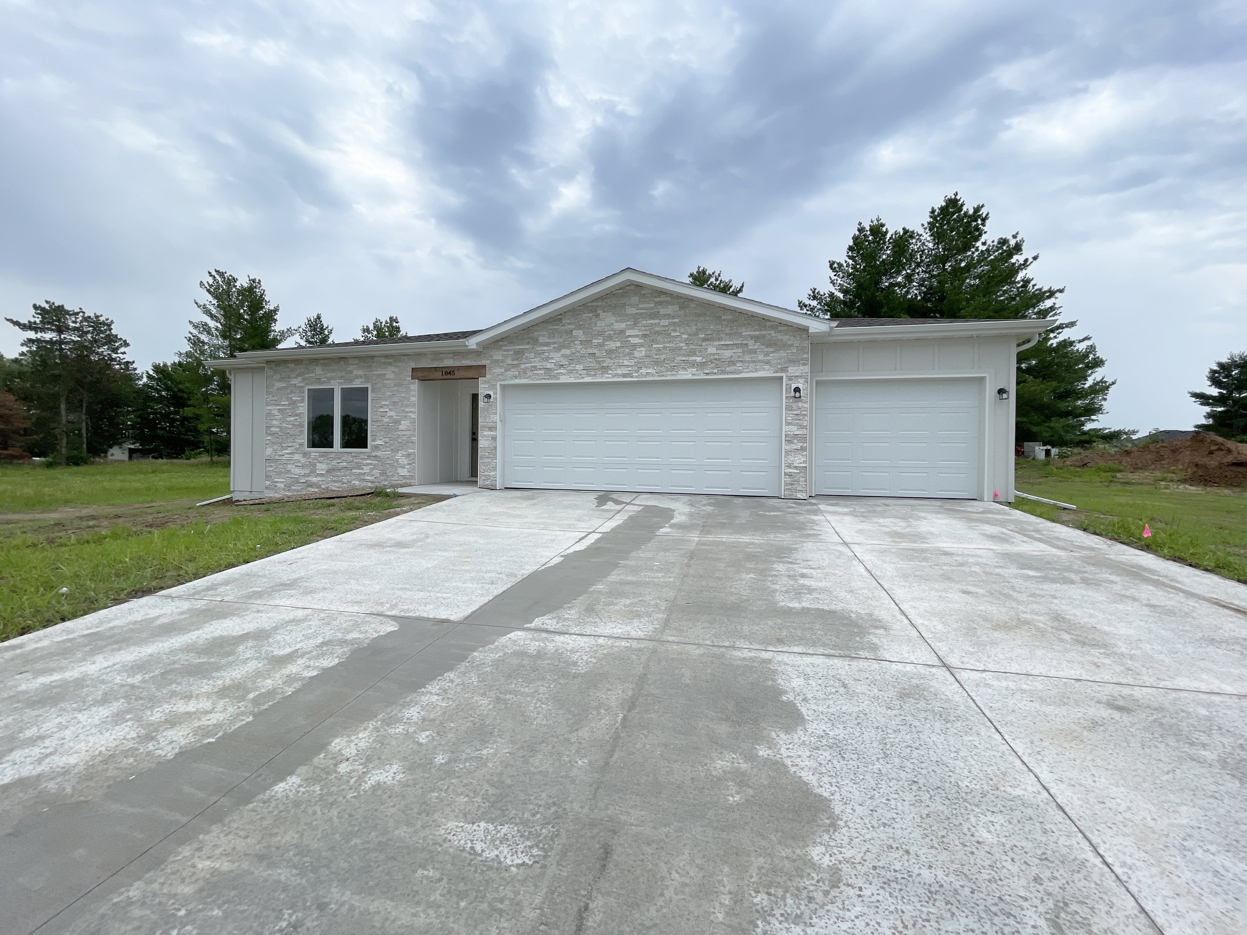 Newly built single-story house with white brick and siding, two-car garage, driveway, and a cloudy sky.