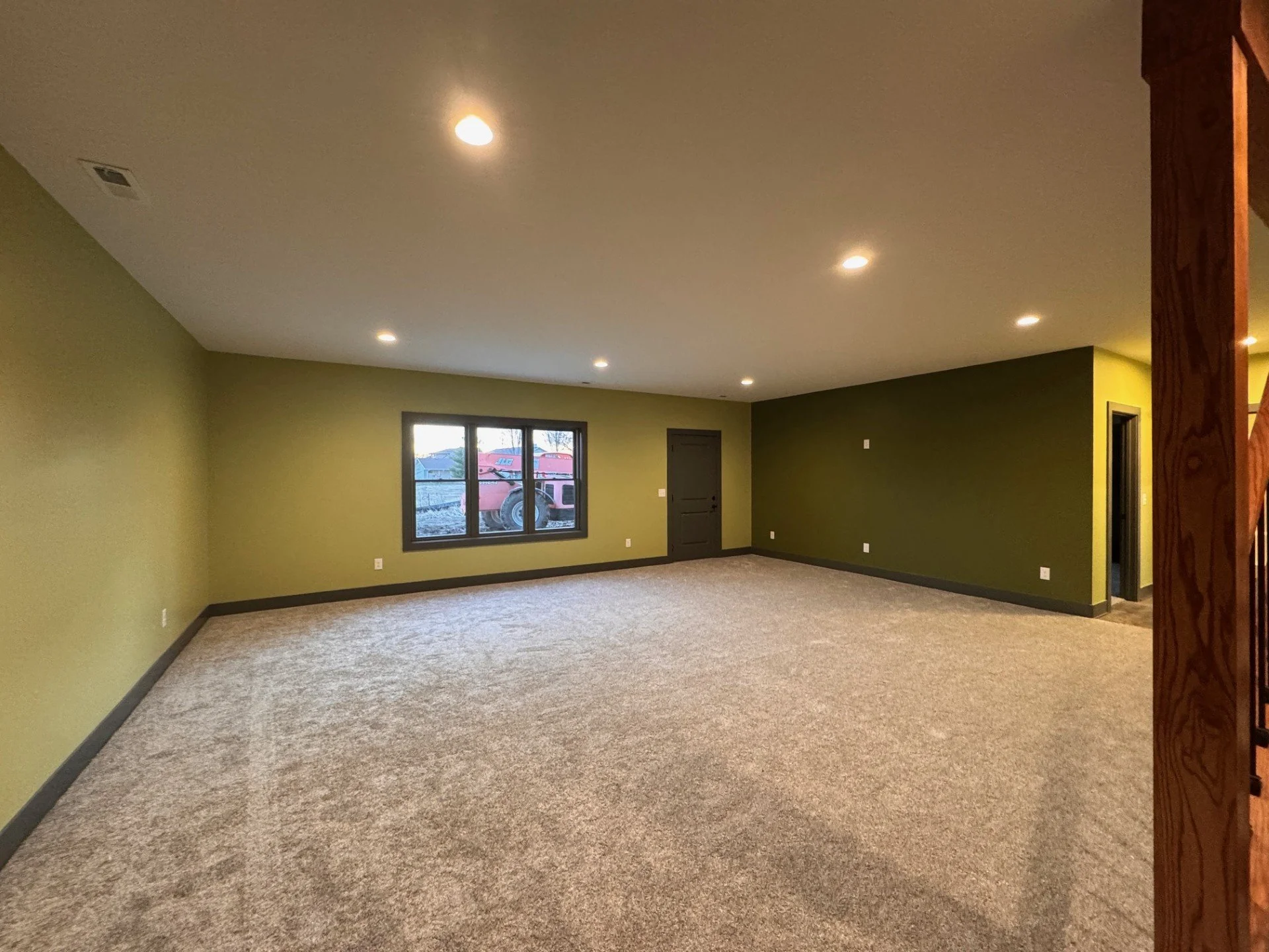 A family room with green walls, large window, beige carpet, and visible ceiling lights.