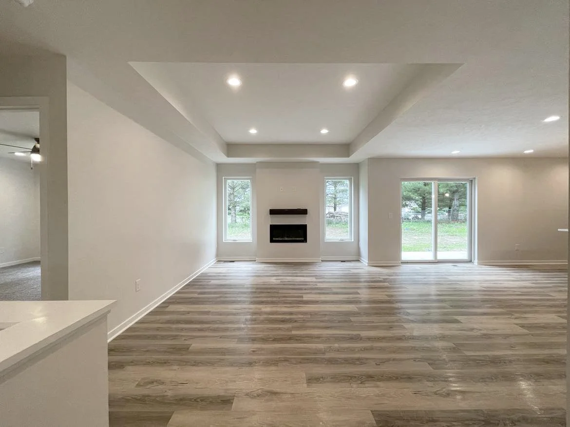 A living room with hardwood floors, white walls, recessed ceiling lights, a fireplace between two windows, and a sliding glass door leading outside.