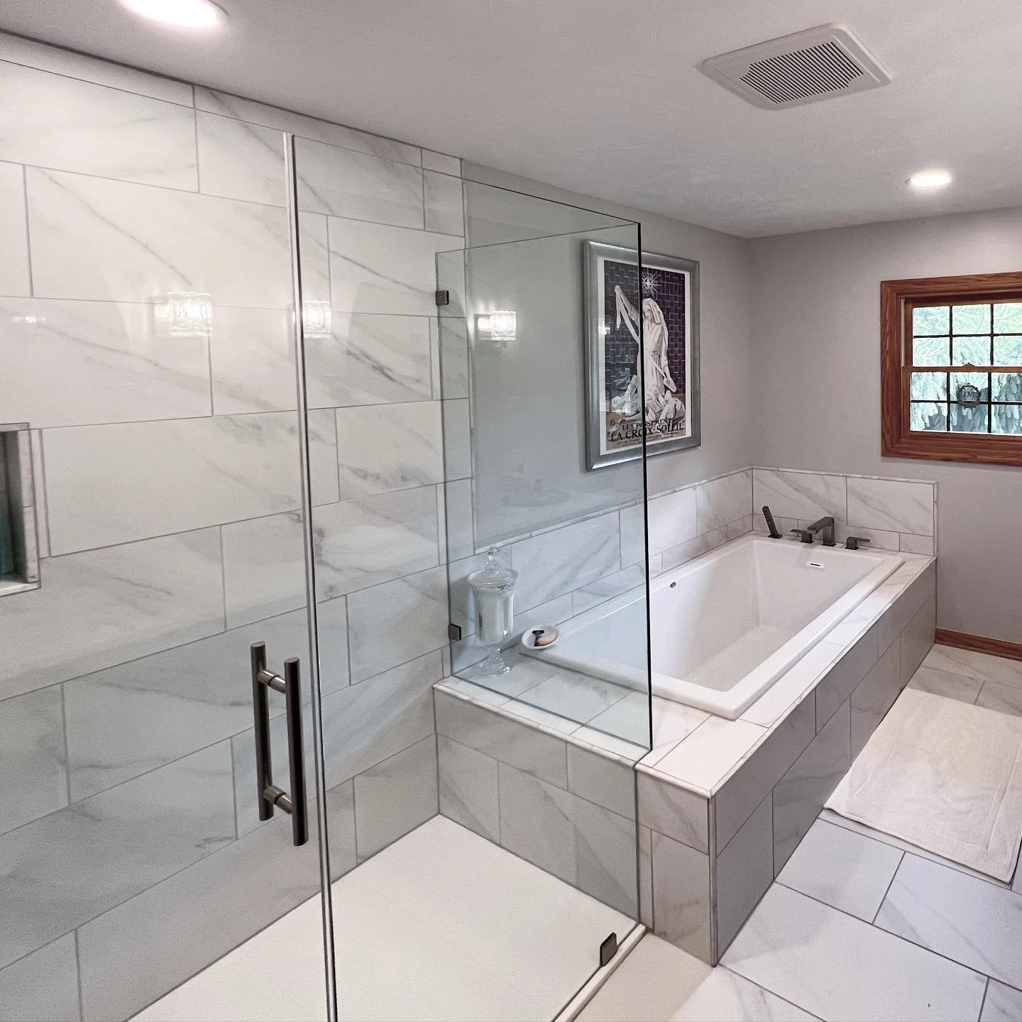 Modern bathroom with a glass shower enclosure, a built-in bathtub, a framed artwork on the wall, a window with wood trim, and a white bath mat on the tile floor.