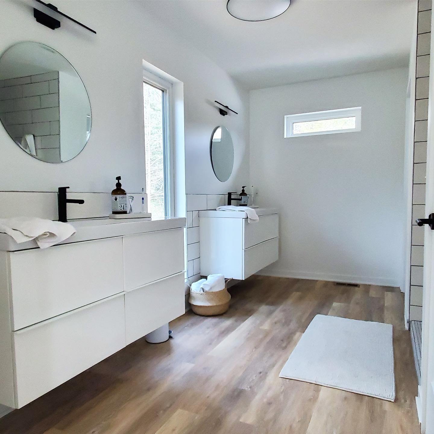 A modern bathroom with white walls, wooden flooring, two floating vanities with black faucets, oval mirrors, a small window above one vanity, a rectangular window on the far wall, a white rug on the floor, and a basket with towels near the vanities.