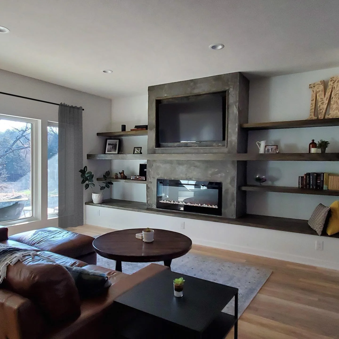 Modern living room with a brown leather sectional sofa, round wooden coffee table, black side table with a potted plant, sliding glass door with gray curtains, built-in shelf with plants and decor, mounted TV above a double-sided fireplace, hardwood floors, and light-colored walls.