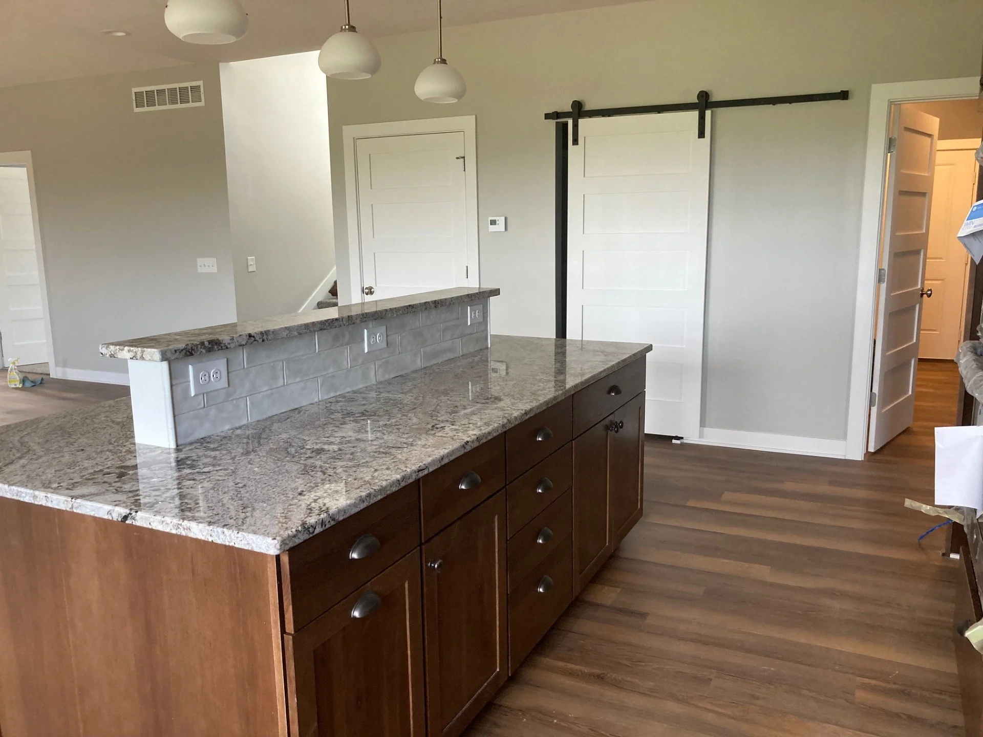 Kitchen with brown cabinets, granite countertops, white subway tile backsplash, and hardwood floors. There are white pendant lights hanging from the ceiling and a white sliding barn door on the wall.