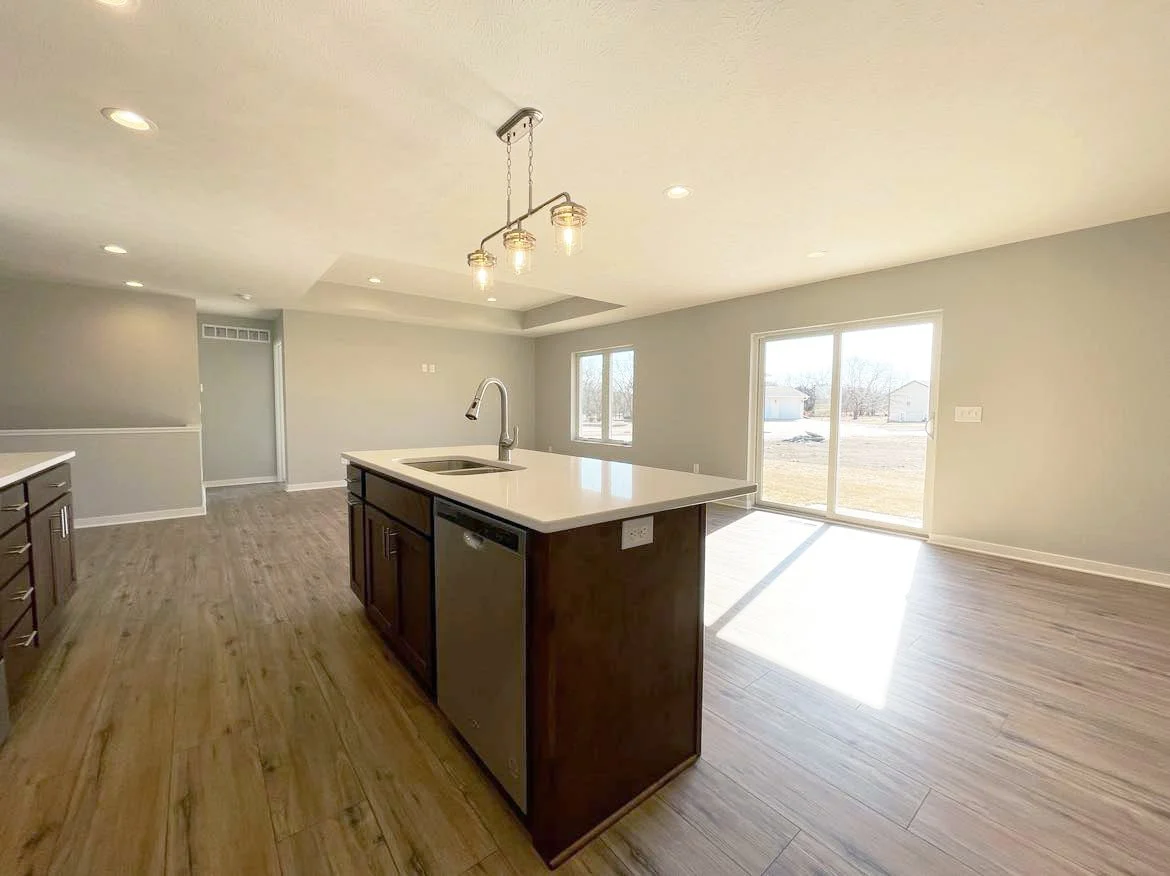 An open-concept kitchen and living area with hardwood floors, light gray walls, large windows, a sliding glass door, and a ceiling light fixture.