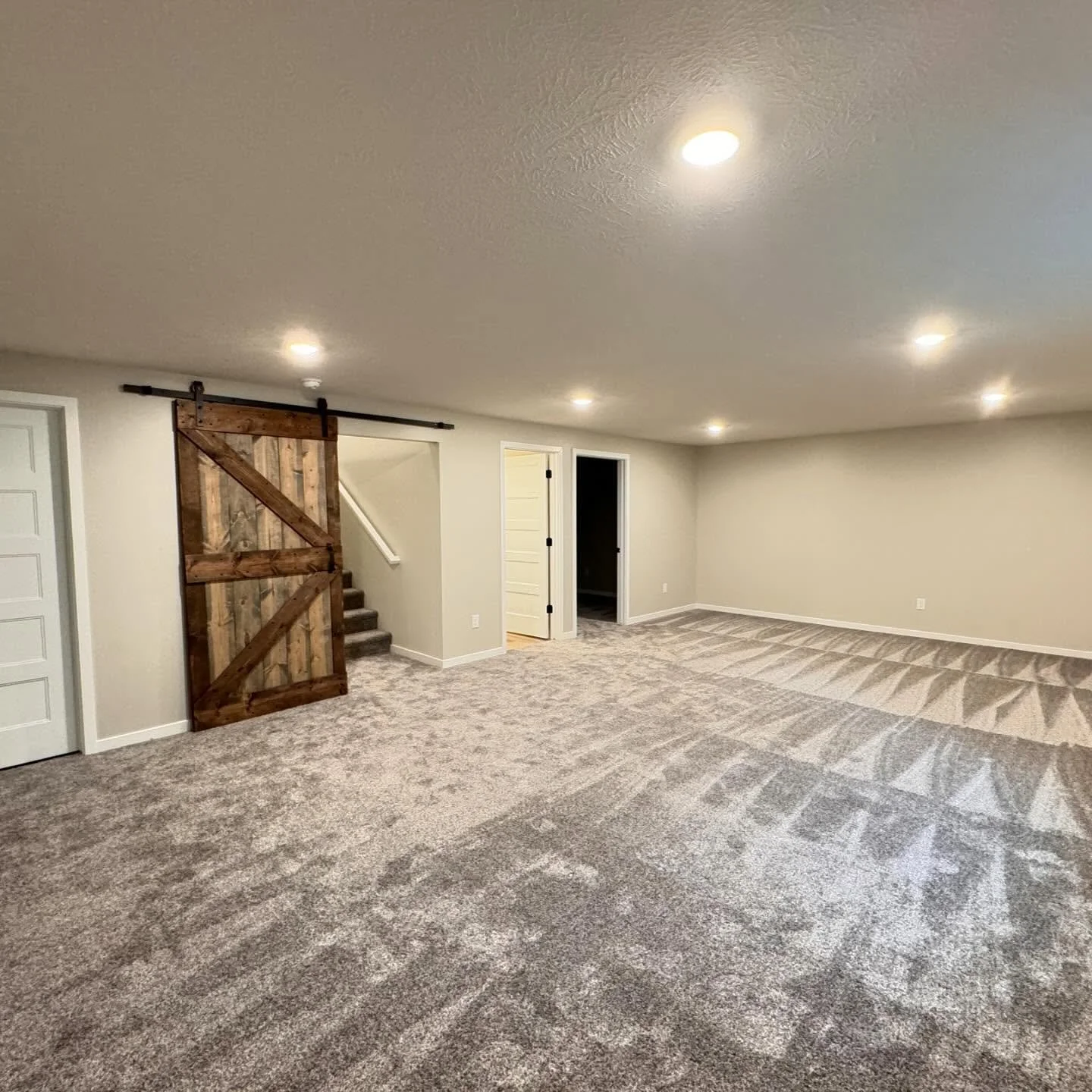 A basement room with beige walls, carpeted floor, recessed ceiling lights, sliding barn door on a black track, stairs behind the door, and open door leading to another room.