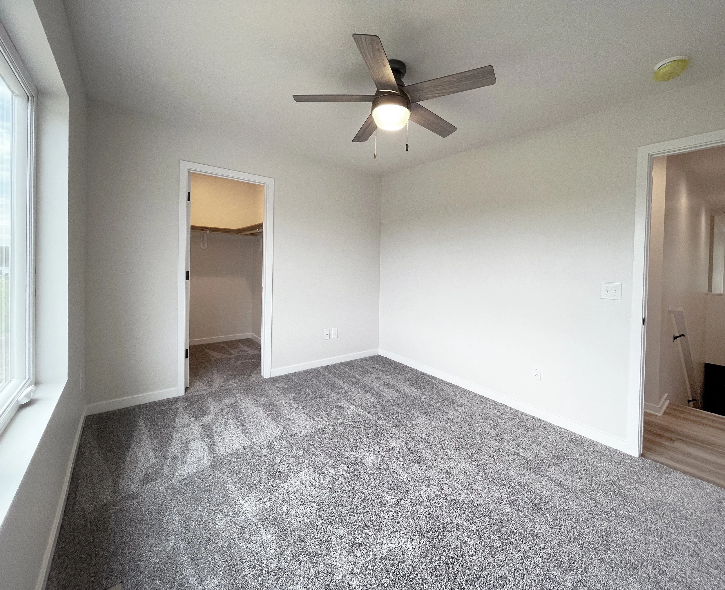 A bedroom with gray carpet, white walls, a window, ceiling fan, and open closet door.