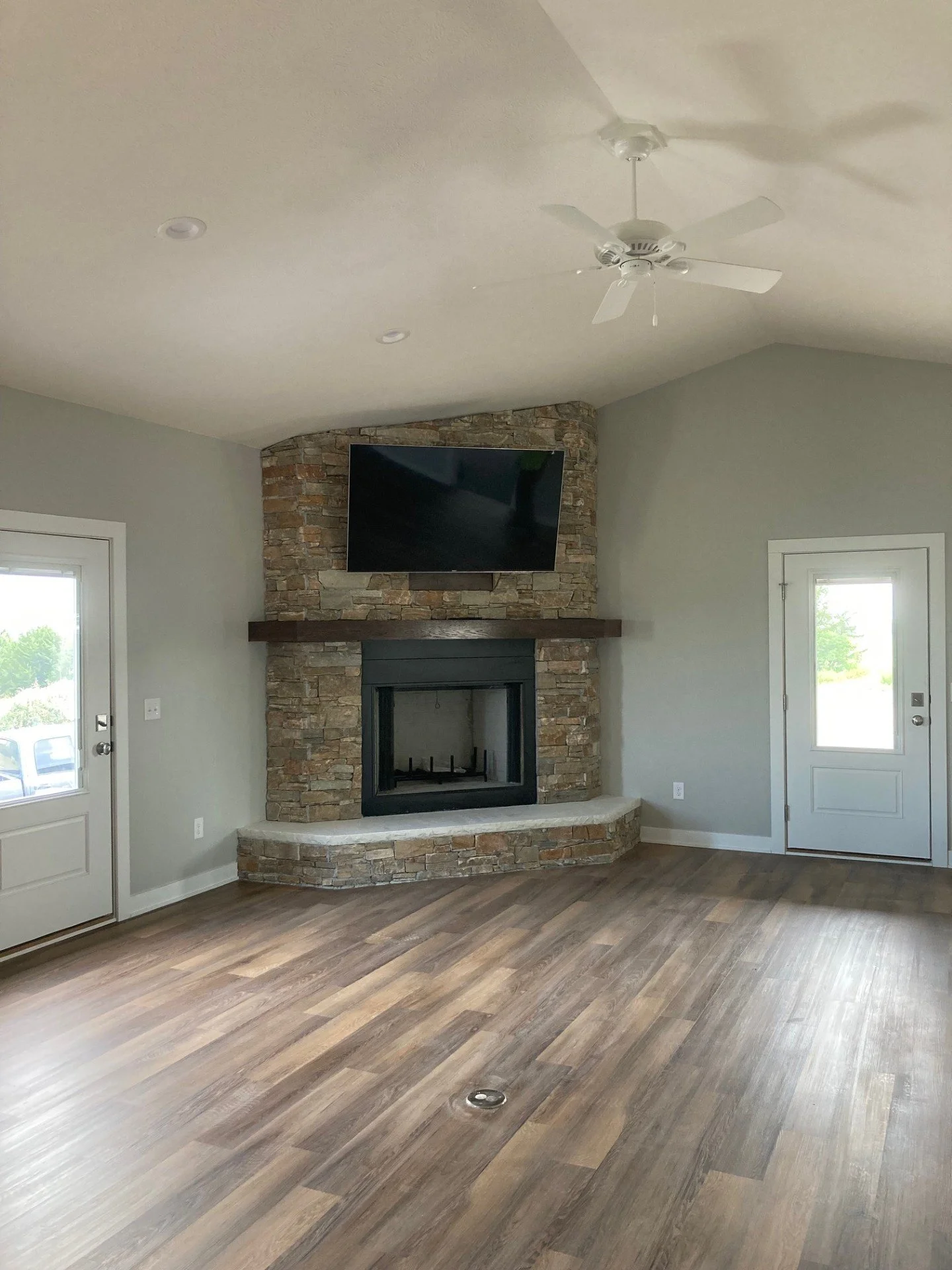 A living room with a stone fireplace and a wall-mounted TV, light gray walls, wood flooring, two white doors with windows, and a ceiling fan.