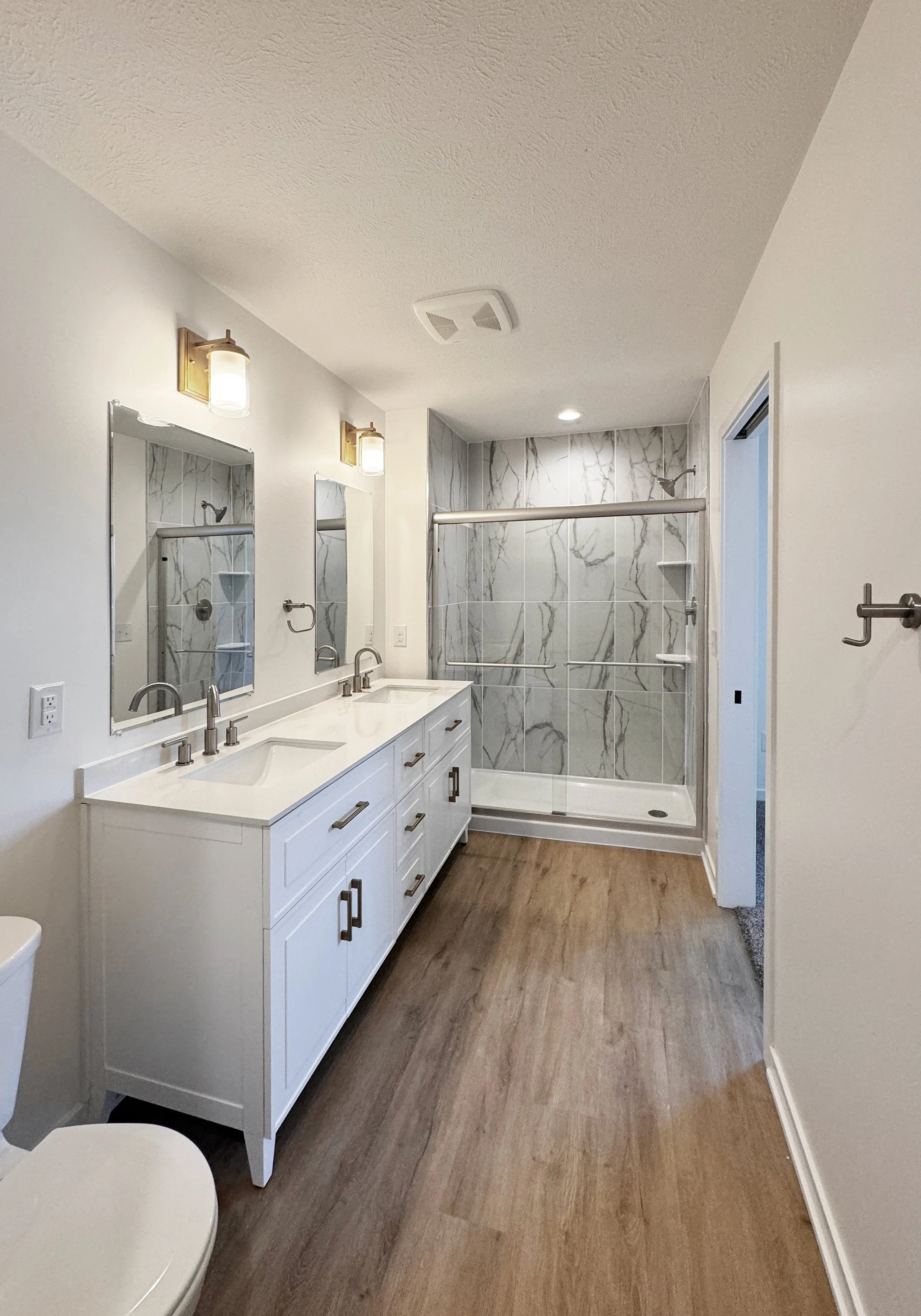 Modern bathroom with dual sinks, white vanity, large mirrors, brass wall lights, walk-in shower with glass door and marble tile walls, and hardwood floor.