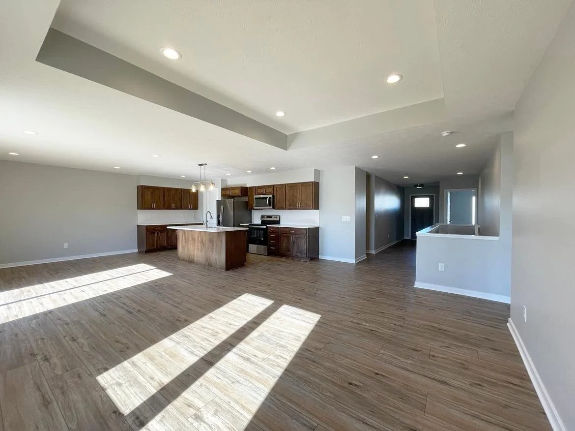 A modern open-concept living and kitchen area with hardwood floors, white walls, and recessed lighting, featuring wooden kitchen cabinets and stainless steel appliances.