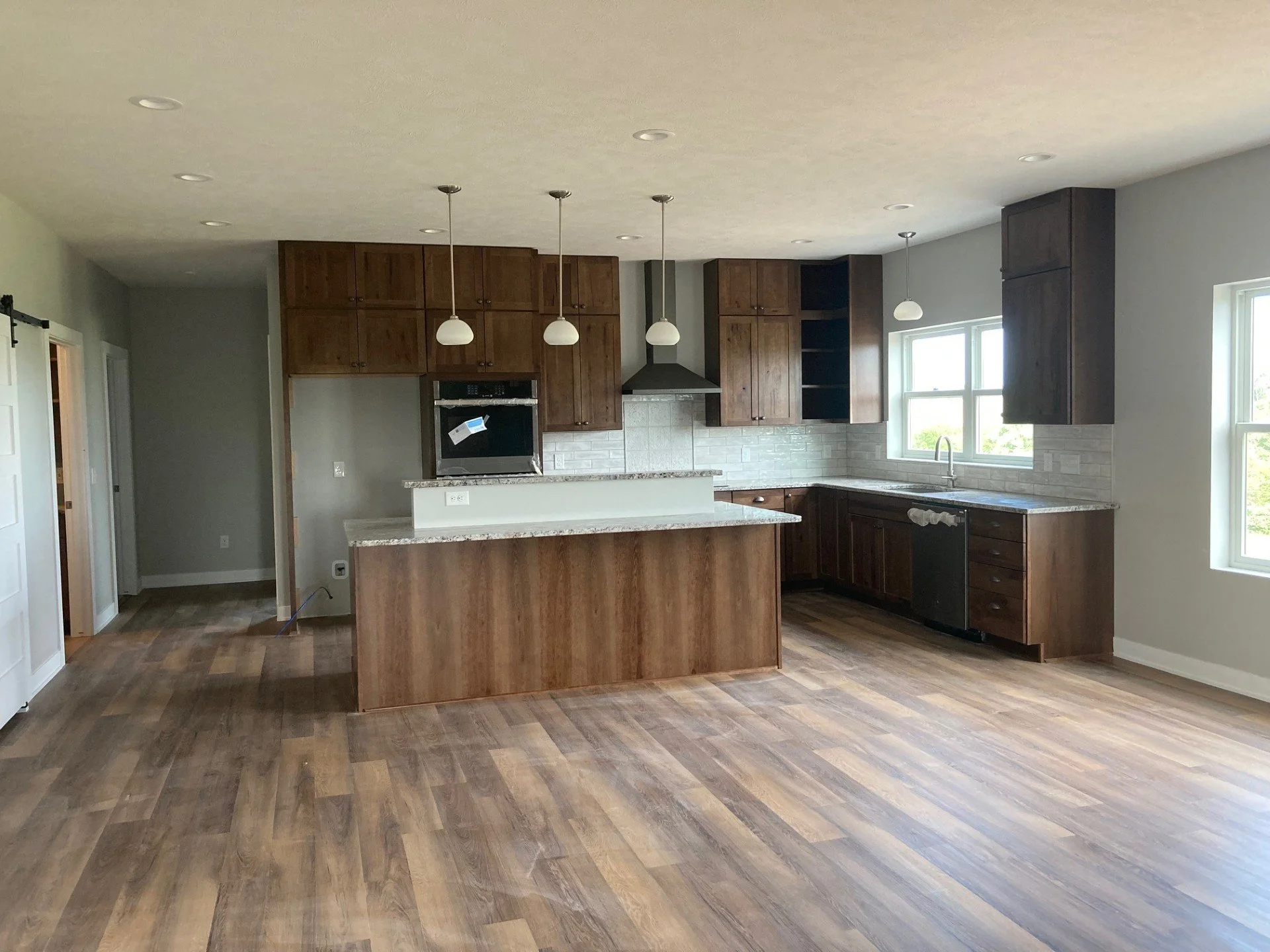 A kitchen with wooden cabinets, granite countertops, and hardwood flooring. There are three pendant lights above the island, large windows letting in natural light, and a stove with a range hood.