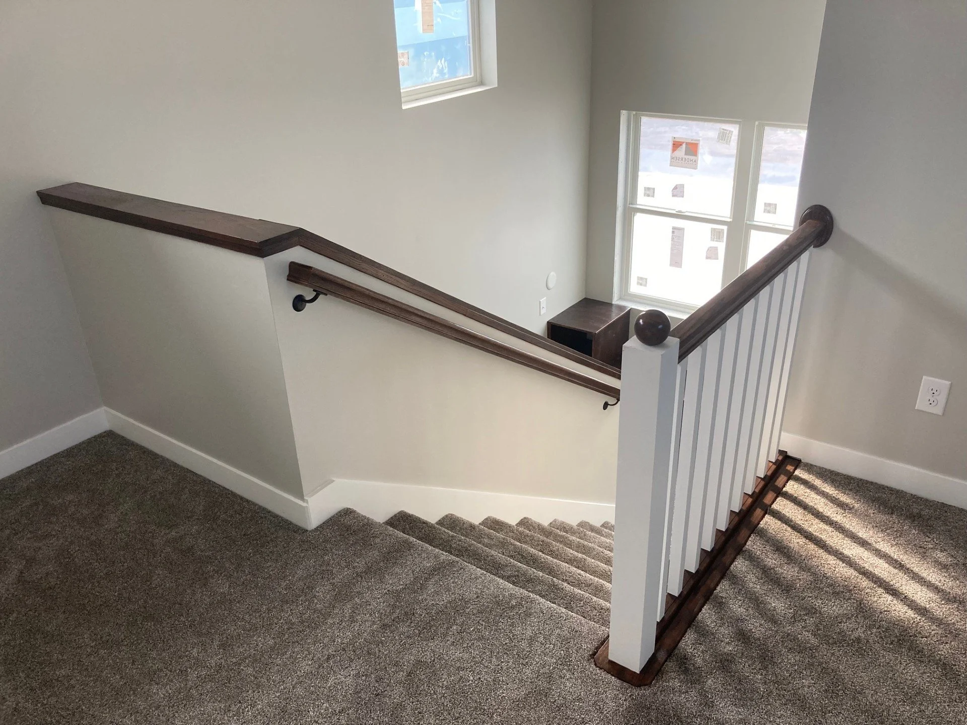 Interior of a staircase landing with carpeting, white walls, and wooden handrails, overlooking a living room area with windows and signs.
