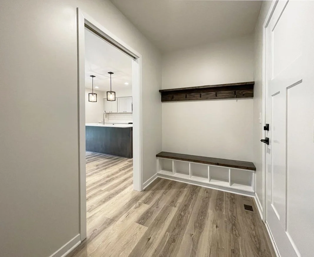 An entryway with wood flooring, white walls, a built-in bench with cubbies, a wall-mounted wooden coat rack with hooks, and an open doorway leading to a kitchen with pendant lights and white cabinets.