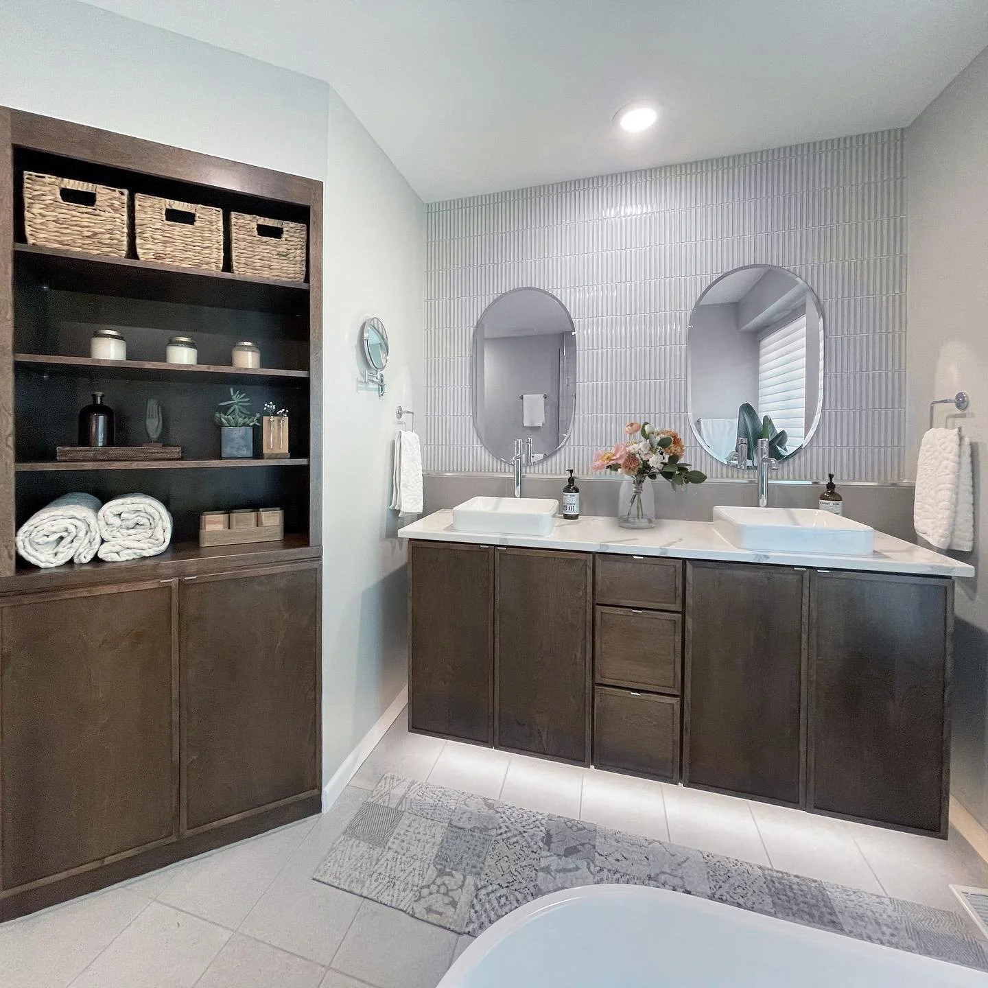 Modern bathroom with double sink vanity, oval mirrors, flower arrangement, and open shelving with towels and decor.
