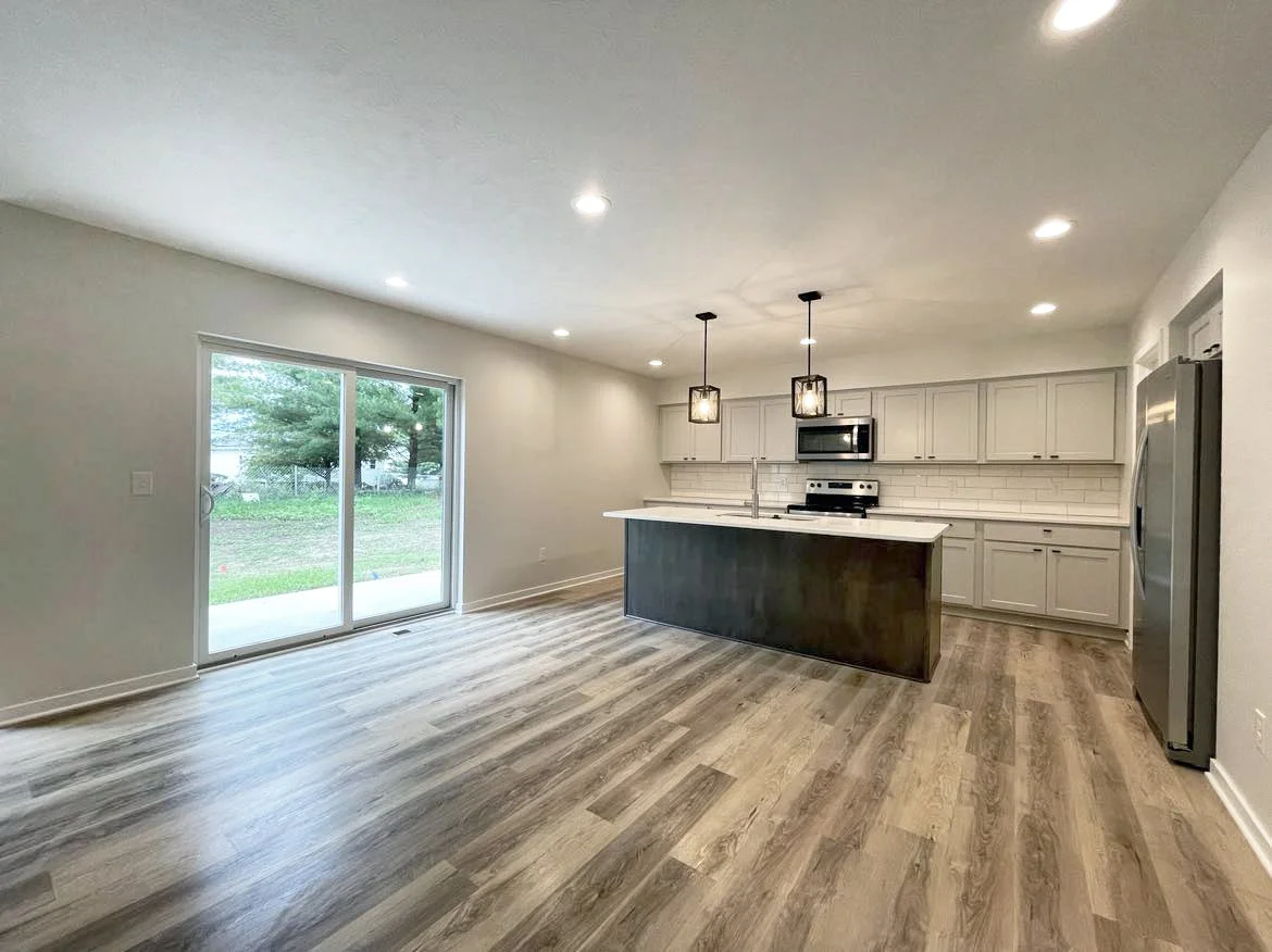 A modern kitchen with white cabinets, stainless steel appliances, a kitchen island with a dark base, pendant lights, sliding glass door, and wood-look flooring.