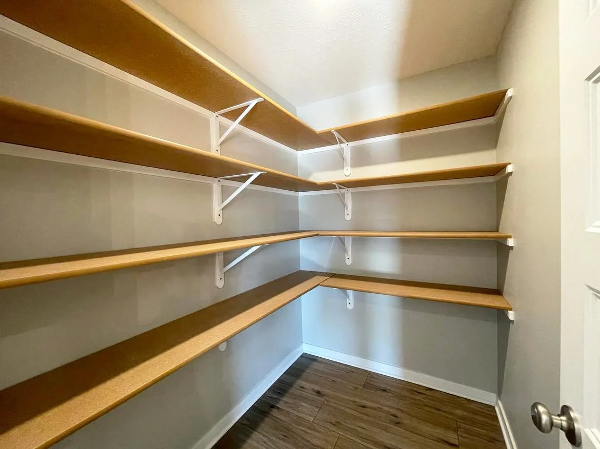 A closet with wooden shelves and white brackets, with hardwood floor and a closed door on the right.
