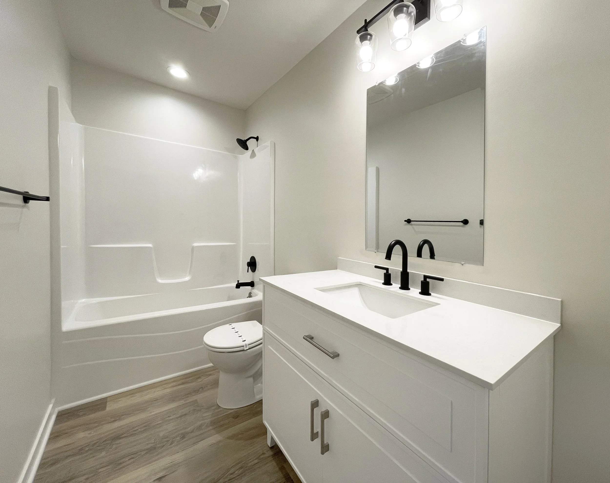 A modern bathroom with a white vanity, black faucet, large mirror, white bathtub with dark fixtures, and wood-look flooring.