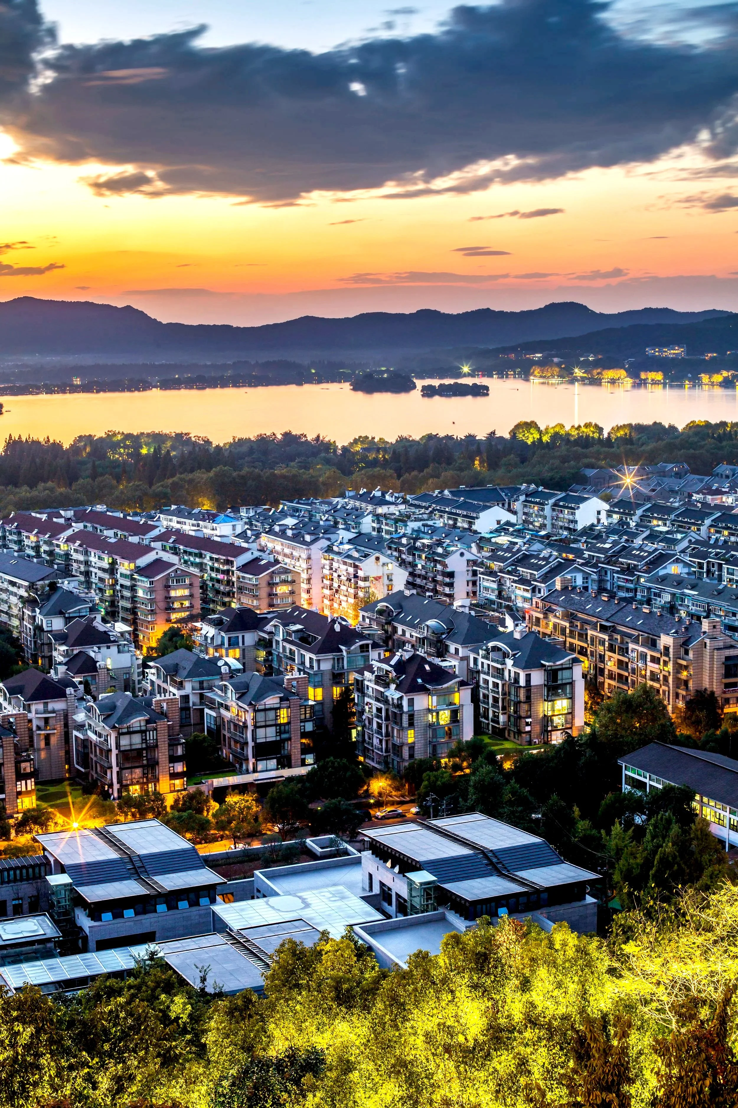 A cityscape at sunset showing modern apartment buildings amidst green trees, with a river and mountains in the background under a partly cloudy sky.