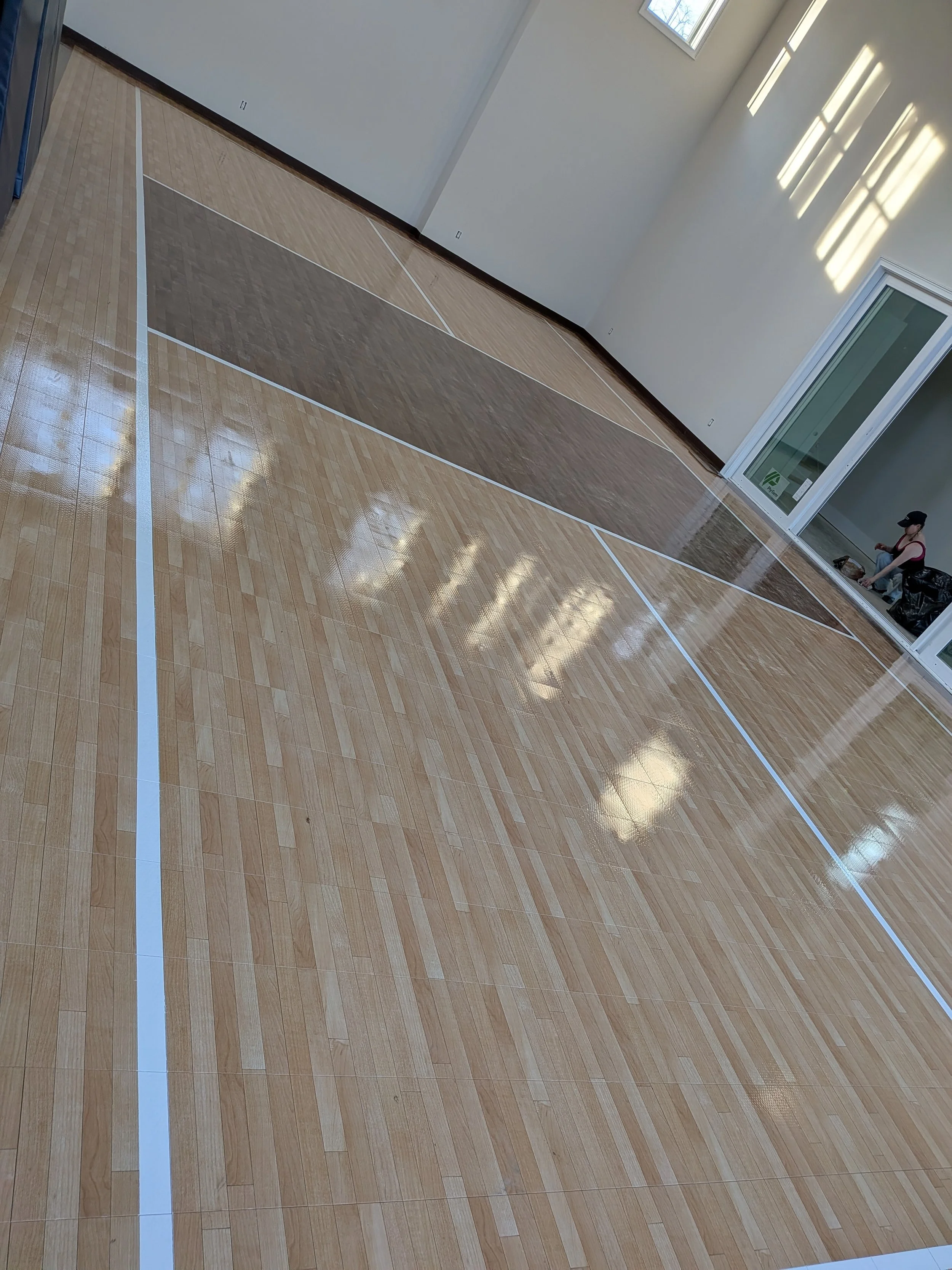 Indoor gymnasium with wooden flooring marked for multiple sports. A person is sitting near a sliding glass door on the right side.