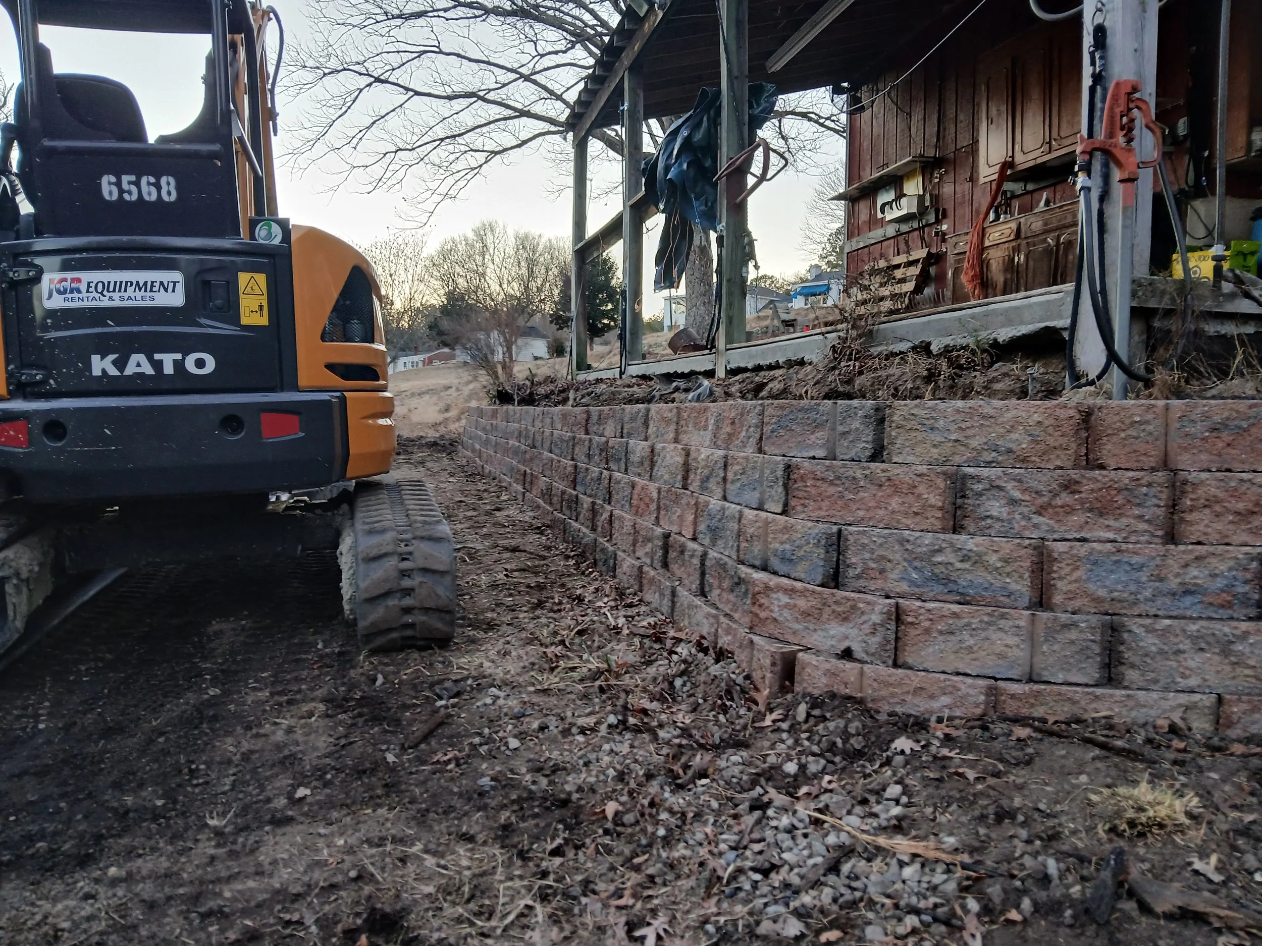Construction site featuring a small excavator near a newly built brick retaining wall, with a wooden shed and tools in the background.