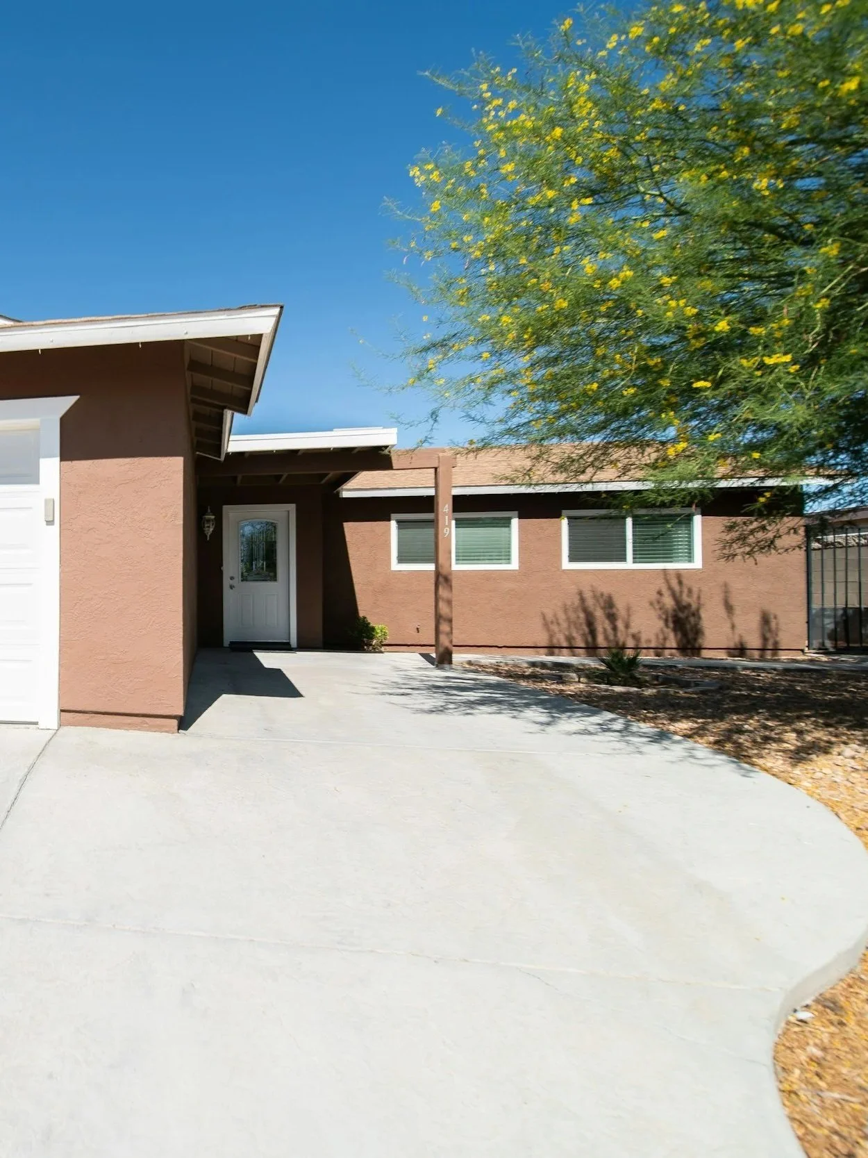 Front view of a single-story house with a brown exterior, white front door, large windows, and a concrete driveway under a clear blue sky. There is a tree with yellow flowers on the right side.