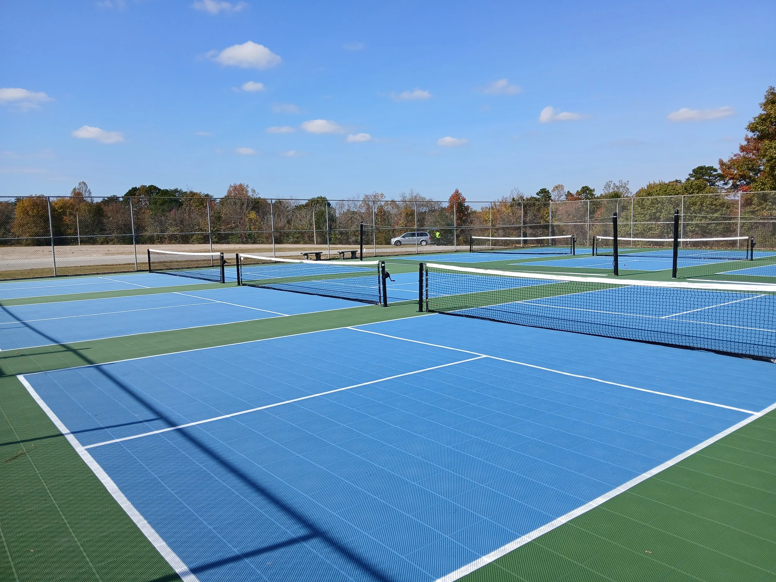 Multiple blue and green outdoor tennis courts with nets, surrounded by a chain-link fence, under a clear blue sky with a few clouds. Trees with autumn foliage are in the background, and a car is parked outside the fence.
