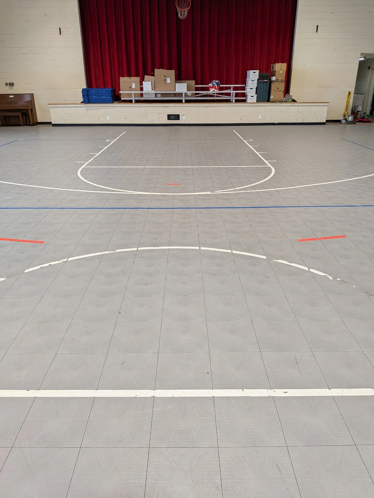 Interior of a gymnasium or auditorium with a basketball court and a stage with a red curtain, surrounded by miscellaneous items and equipment.