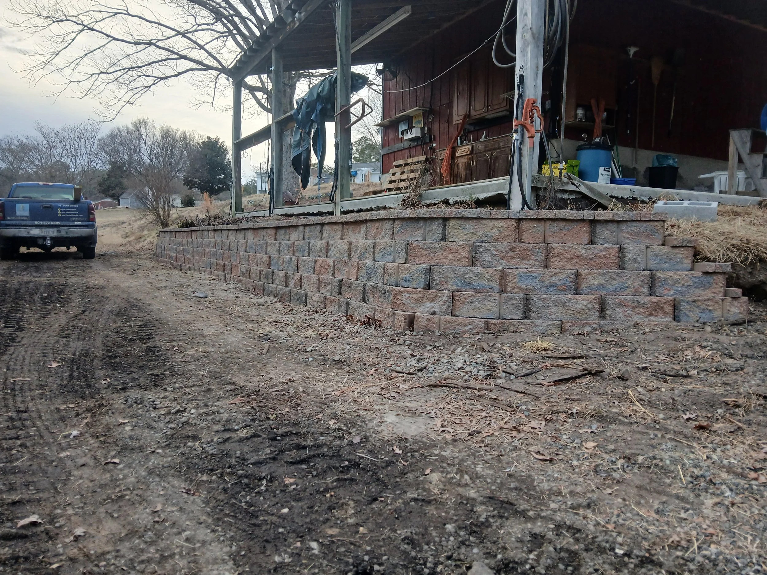A brick retaining wall under construction in front of a house with a porch, with tools and supplies nearby. A pickup truck is parked on a dirt road to the left, and leafless trees are in the background.