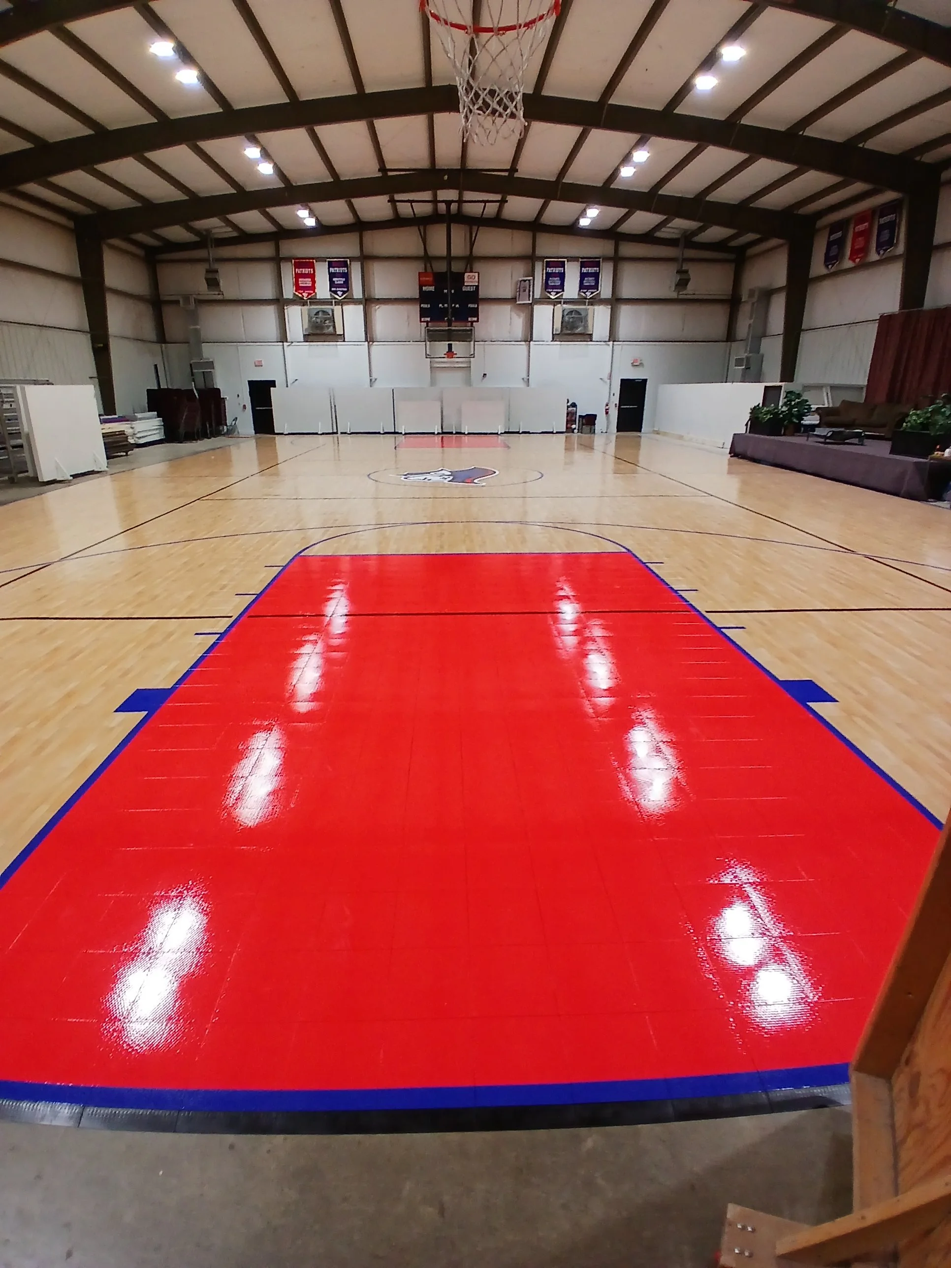 An indoor basketball court with a polished wood floor, marked with red and blue lines, and two basketball hoops at each end. The ceiling is high with bright overhead lights, and there are banners hanging on the back wall.