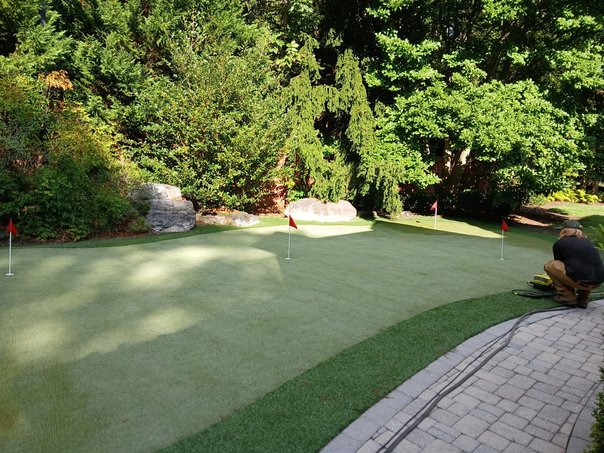 A person crouching on a paved patio next to a miniature golf green, surrounded by lush green trees and bushes, with four small flags marking the holes.