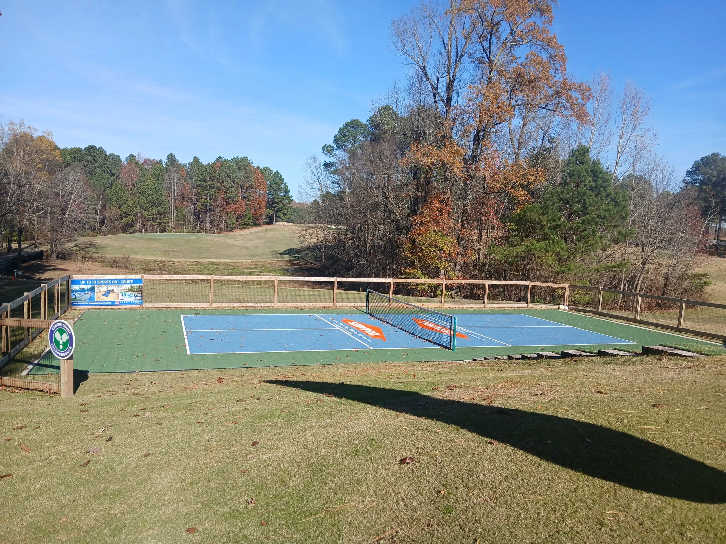 Outdoor tennis court with a blue court surface and a green border, surrounded by a wooden fence, in a park-like setting with trees in the background and a partly cloudy sky.
