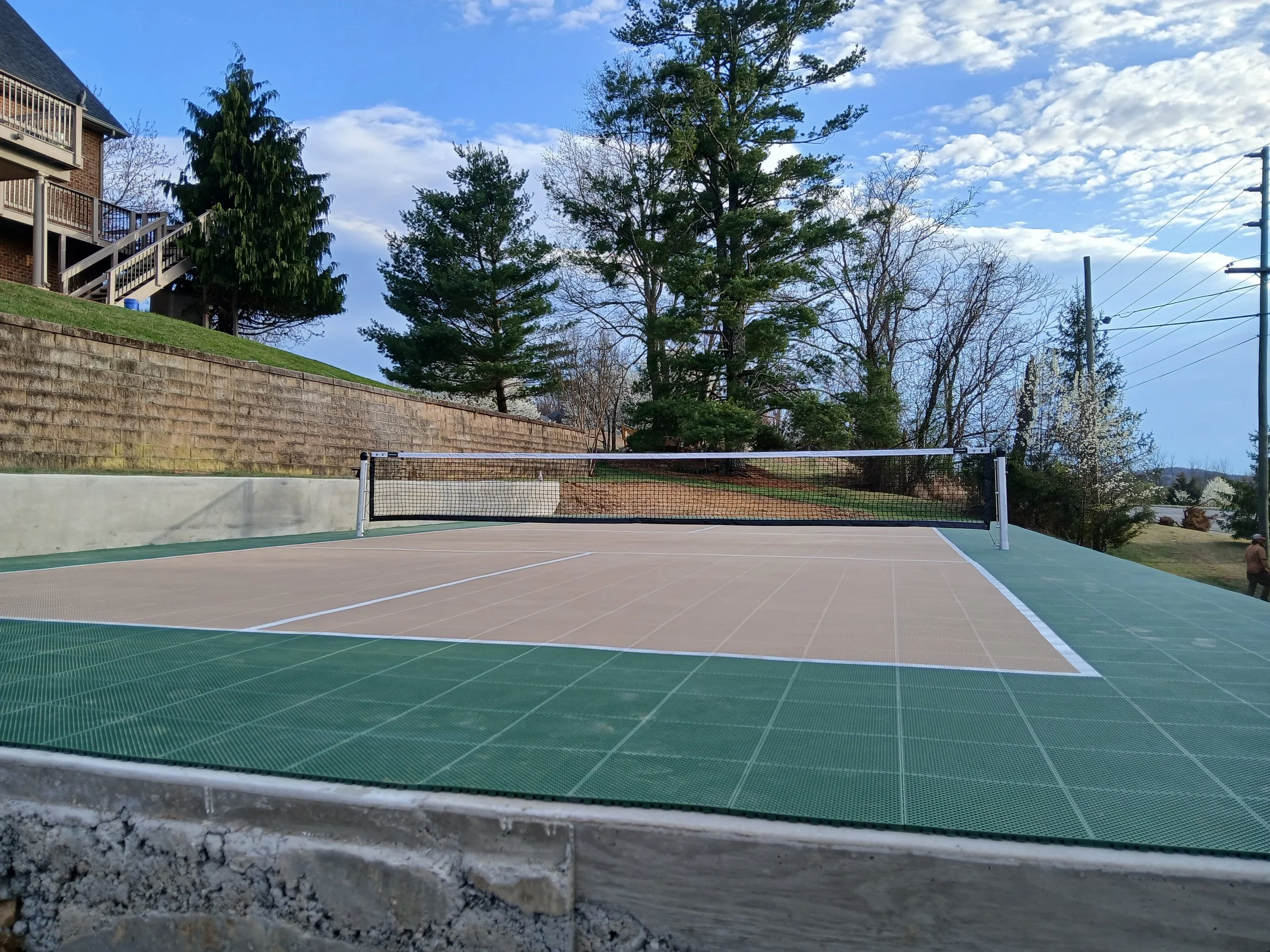 An empty outdoor tennis court on a sunny day, surrounded by trees and residential buildings.