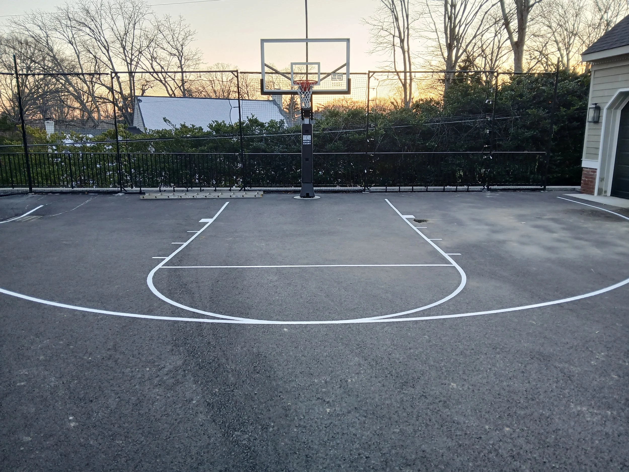 Empty outdoor basketball court at sunset with a hoop, white painted lines, and a privacy fence in the background.