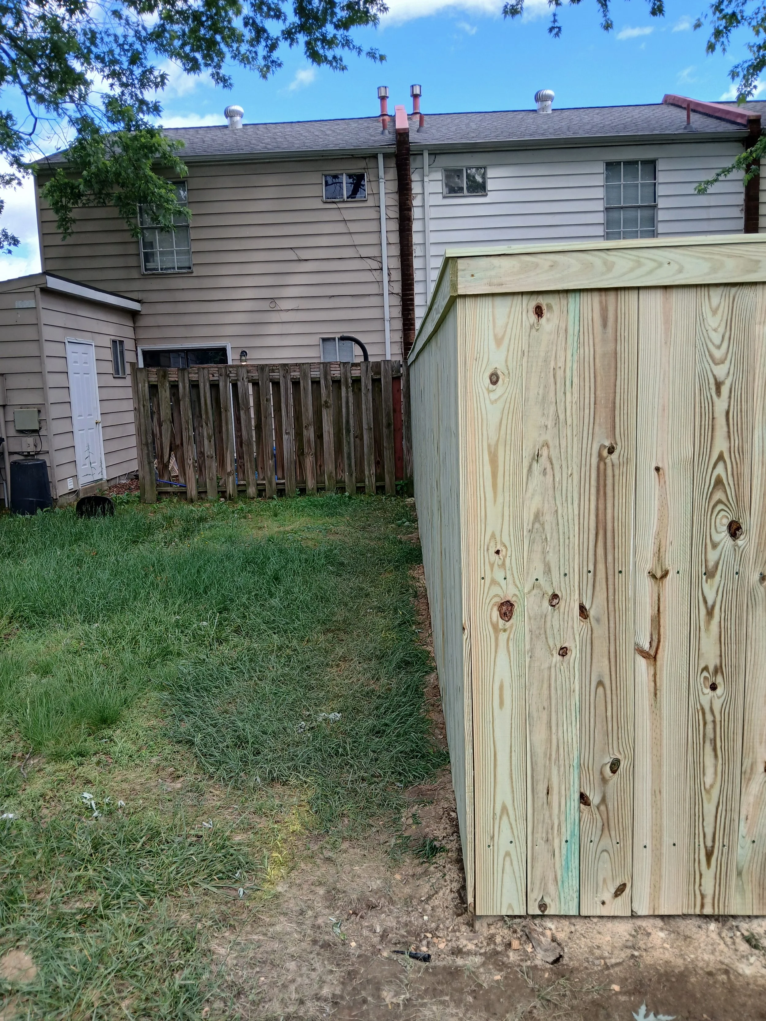 View of a backyard with a new wooden fence on the right side, a grass area, a small shed in the corner, a taller wooden fence in the background, and a two-story house with beige and white siding behind the fences. The sky is partly cloudy.