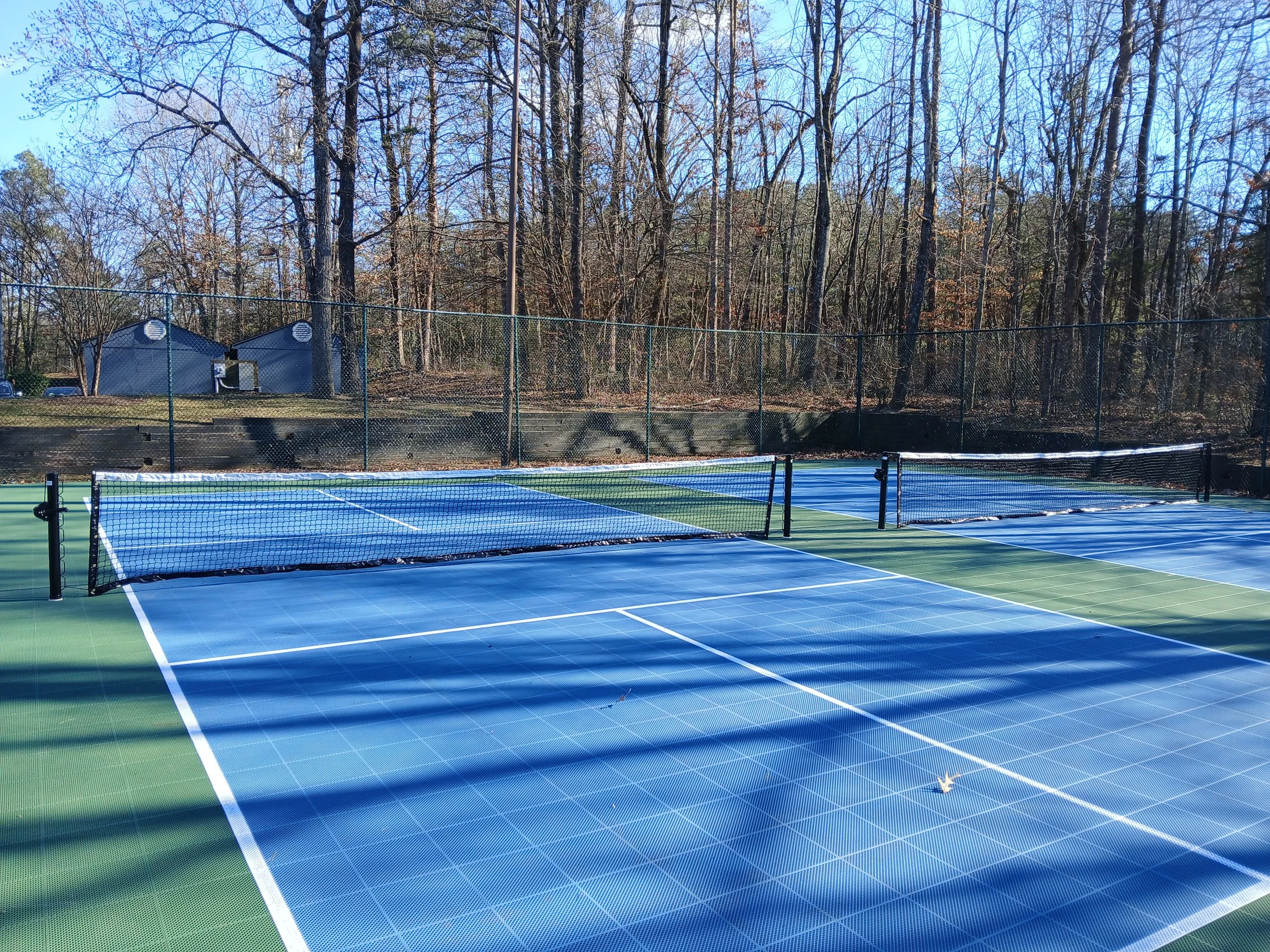 Multiple tennis courts with blue and green surfaces, surrounded by a black chain-link fence, with leafless trees and a couple of storage sheds in the background on a clear, sunny day.