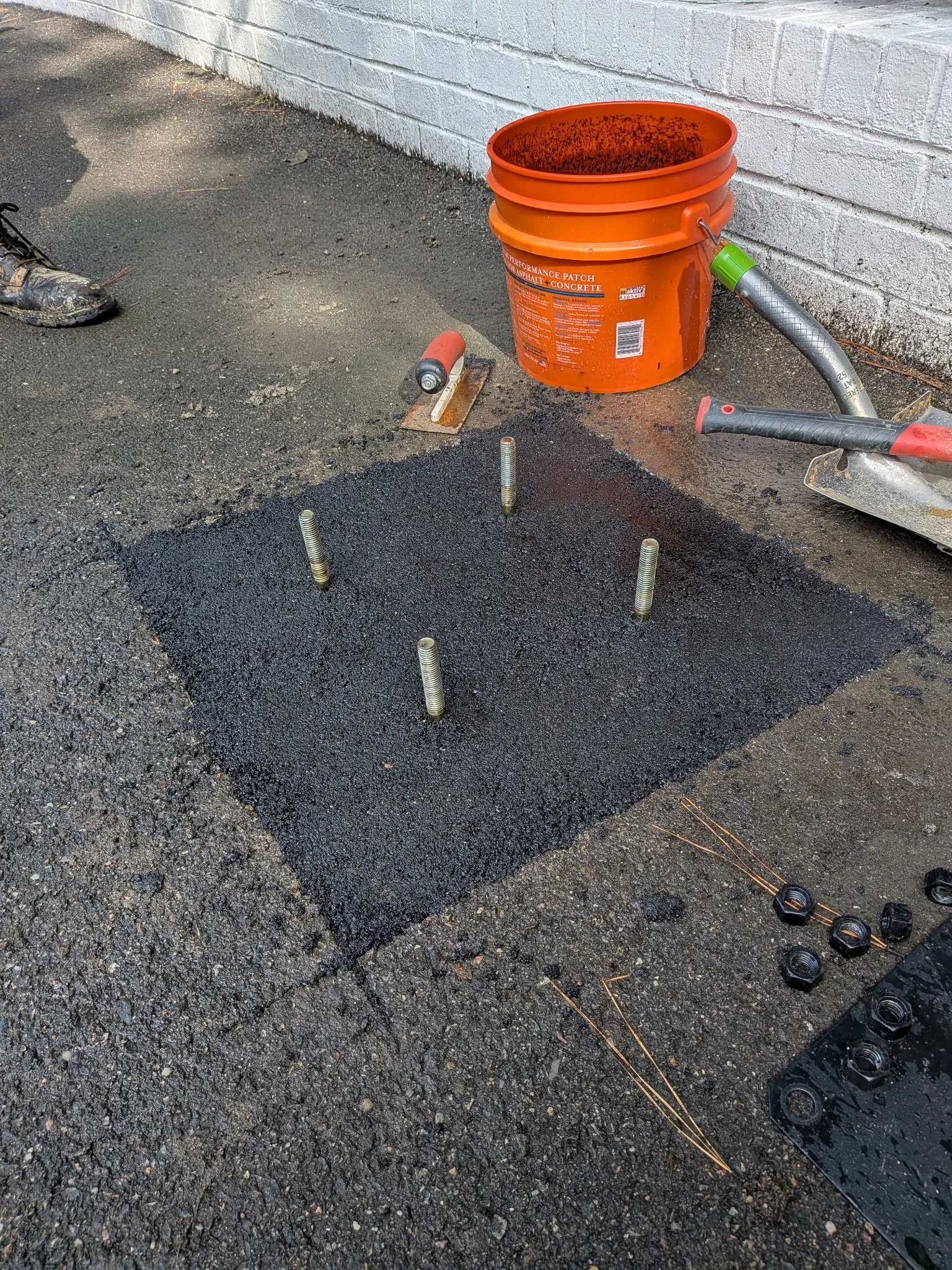 Construction site with a black asphalt surface, concrete wall, and various tools including an orange bucket, a roller, and a trowel. Part of a person's shoe is visible on the left.