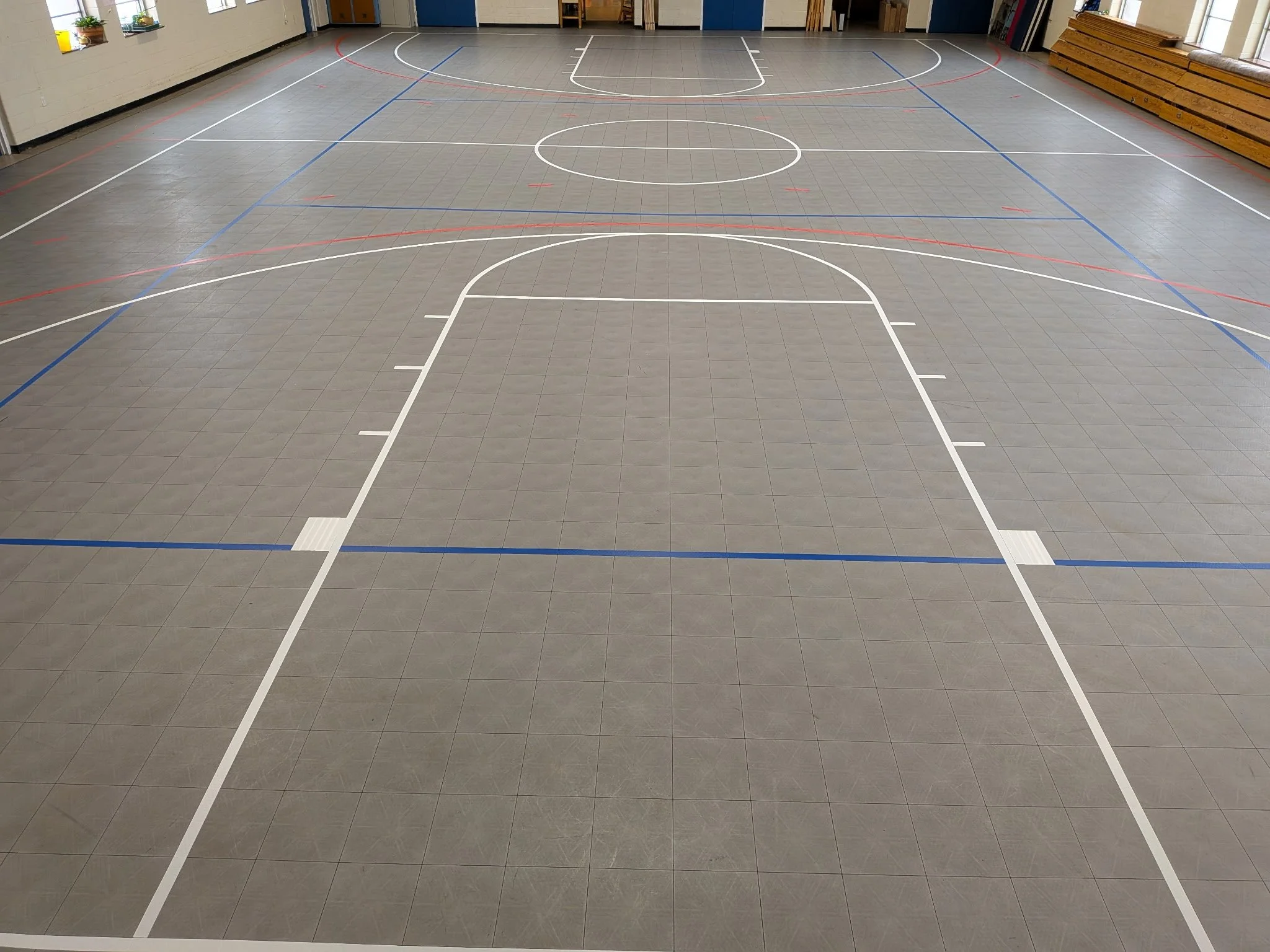 An indoor gymnasium with a gray tiled floor marked with white, blue, and red lines for various sports. The gym is well-lit with windows and has wooden benches along one wall.