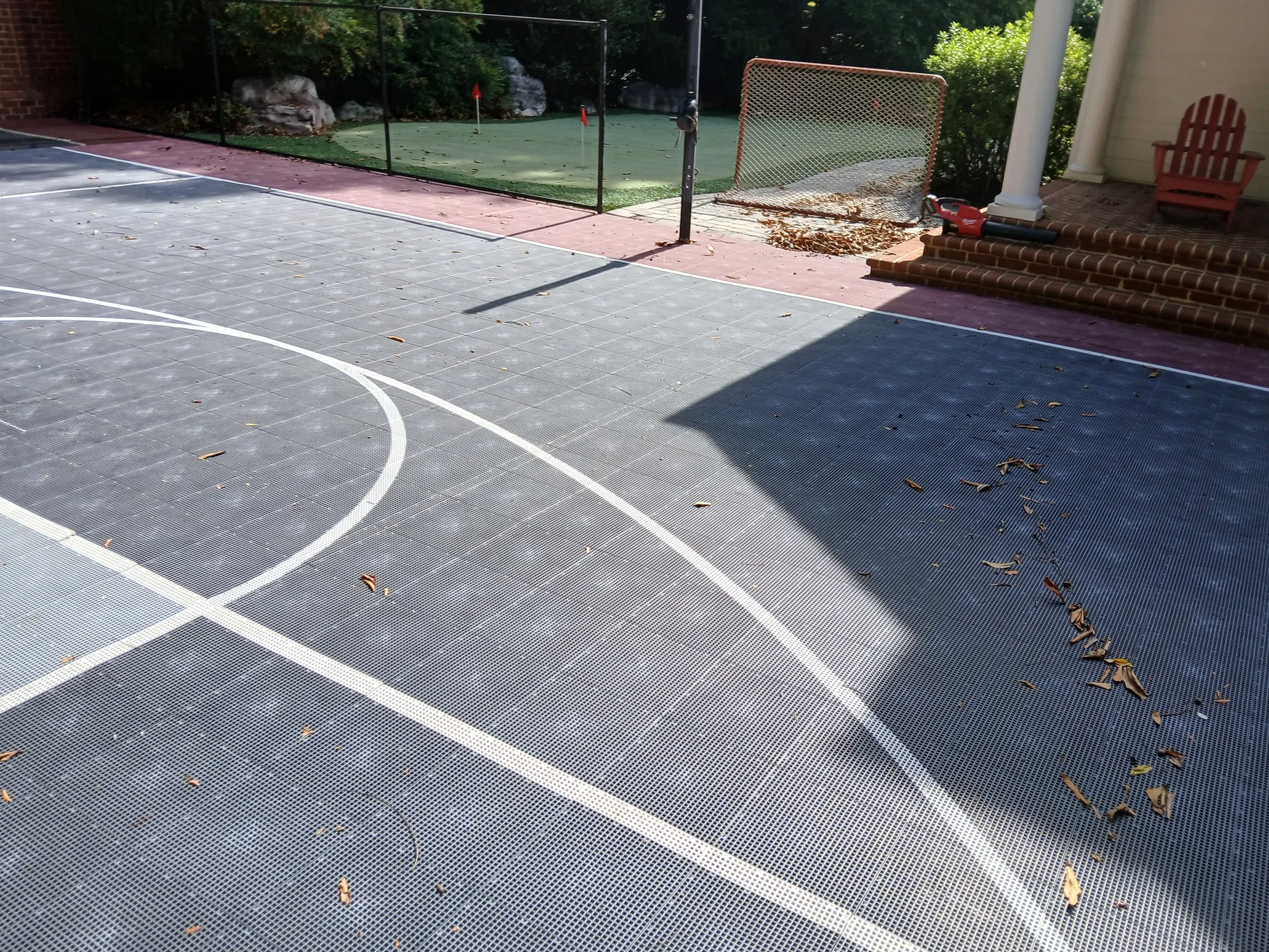 Empty outdoor basketball court with scattered leaves, a green putting area with flags, a brick staircase with a power tool, and a red chair.