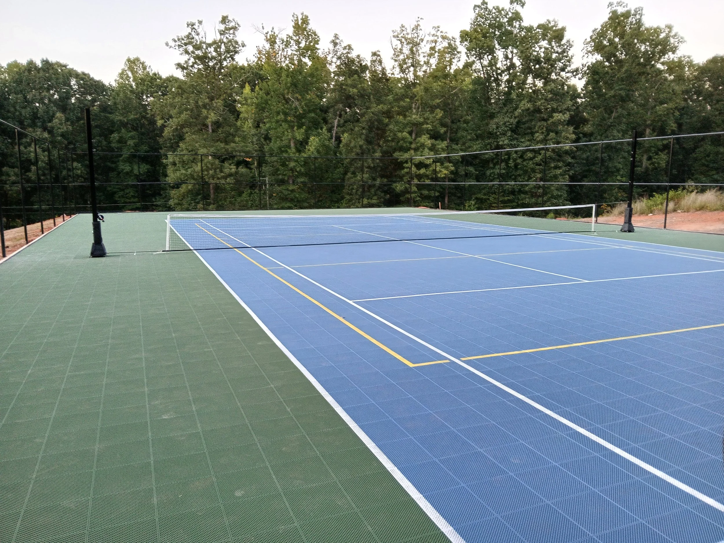An outdoor tennis court with a blue playing surface, surrounded by green flooring and a black fence, with trees in the background.