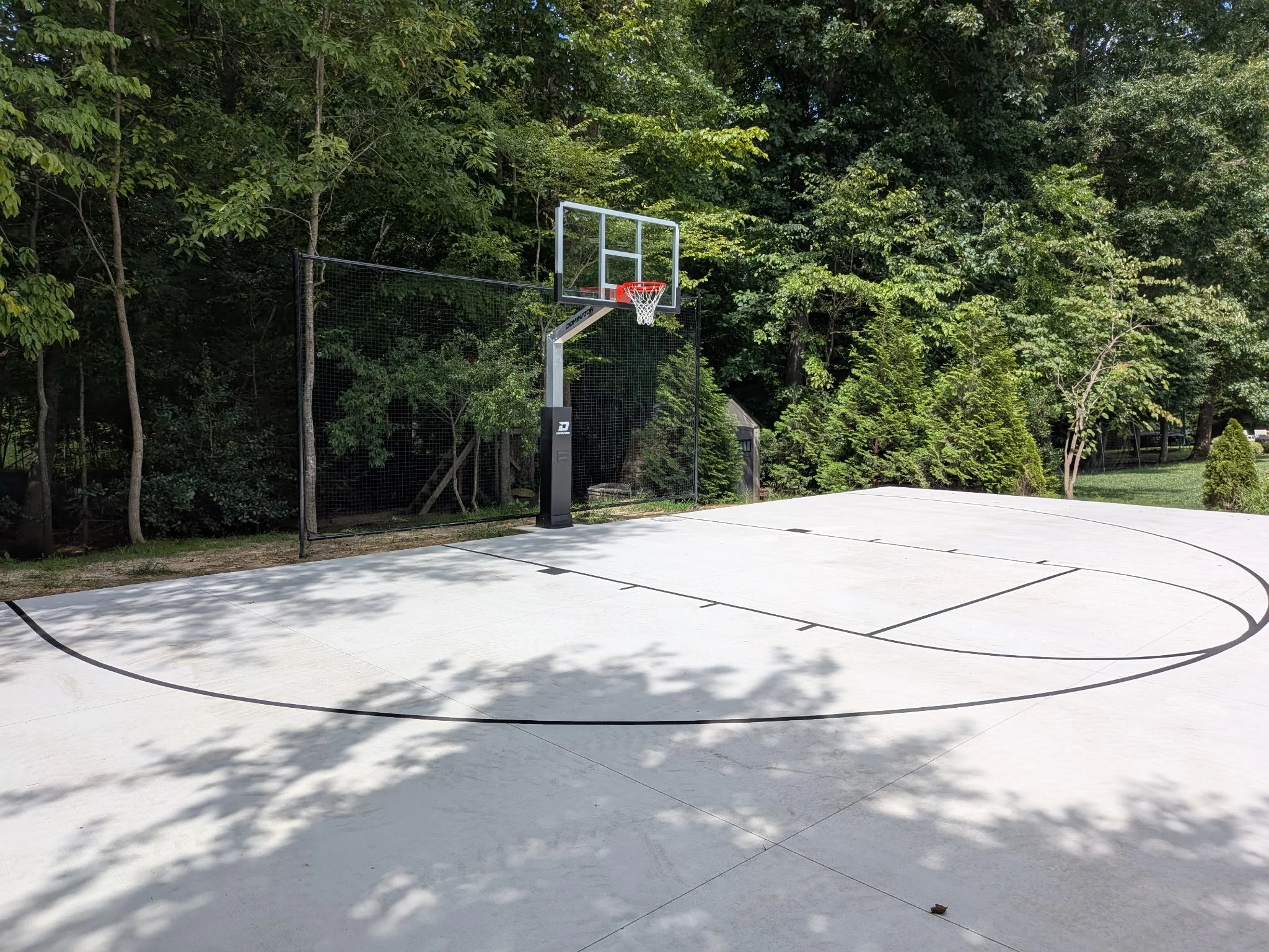 An outdoor basketball court with a basketball hoop and backboard, surrounded by trees and a tall fence.