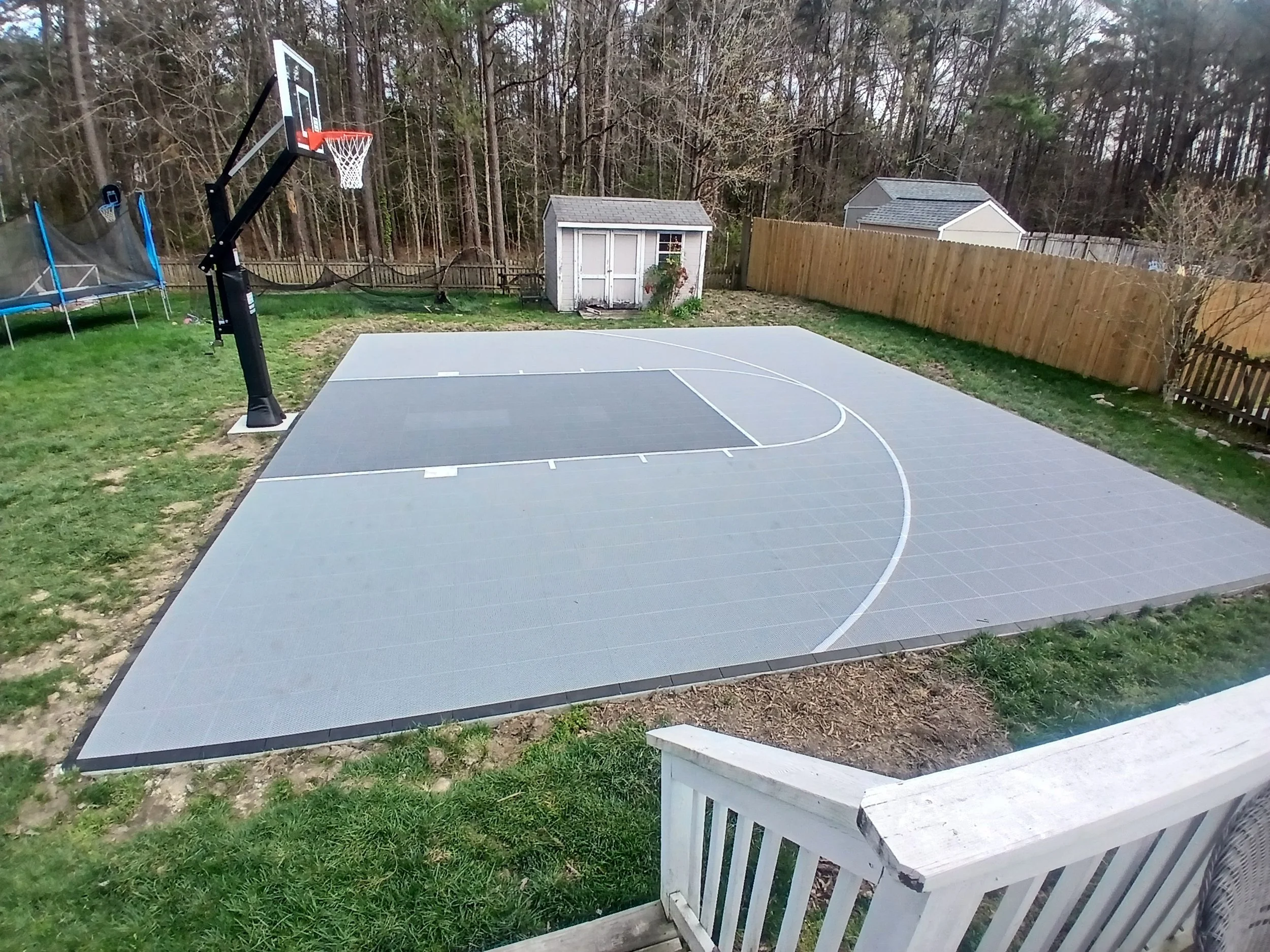An outdoor basketball court with a basketball hoop, surrounded by a backyard with a wooden fence, small sheds, and trees in the background.