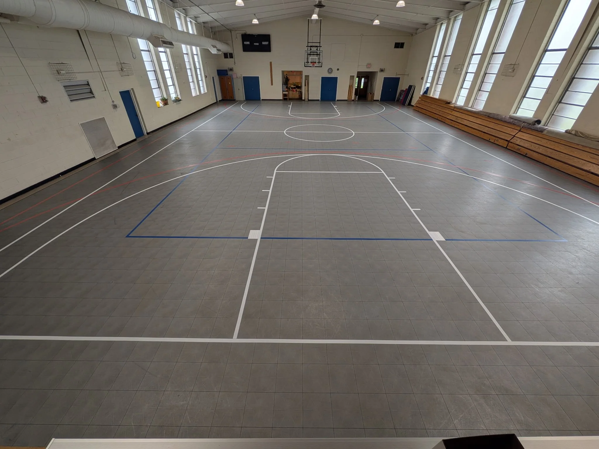 An empty indoor basketball court with white lines marking the playing area. The court has large windows on both sides letting in natural light, and wooden bleachers are stacked along one side. The walls are painted white, and the ceiling has several 
