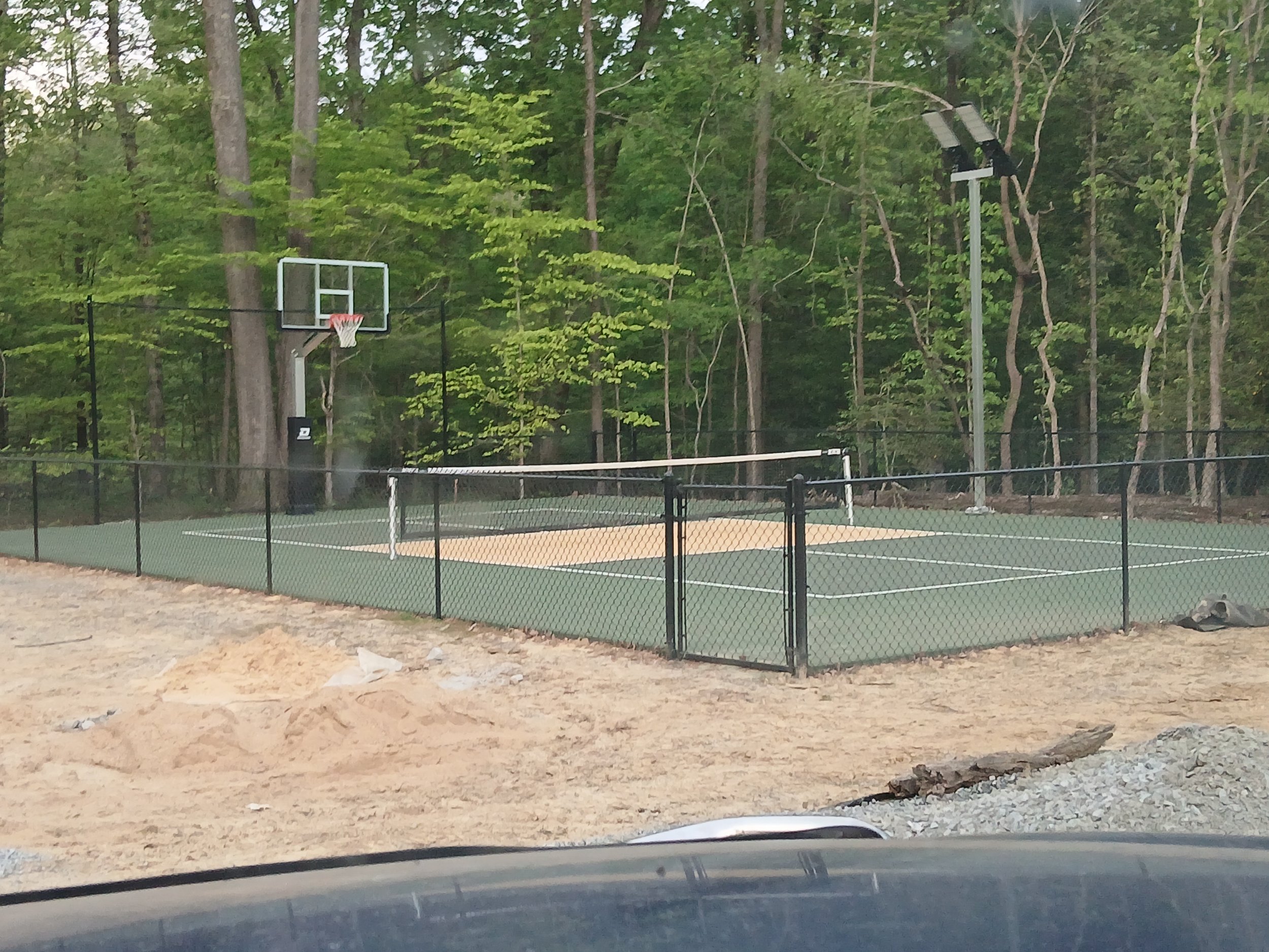 An outdoor basketball court with a basketball hoop and a volleyball net, surrounded by black chain-link fencing and tall trees in the background, with a construction area in the foreground.