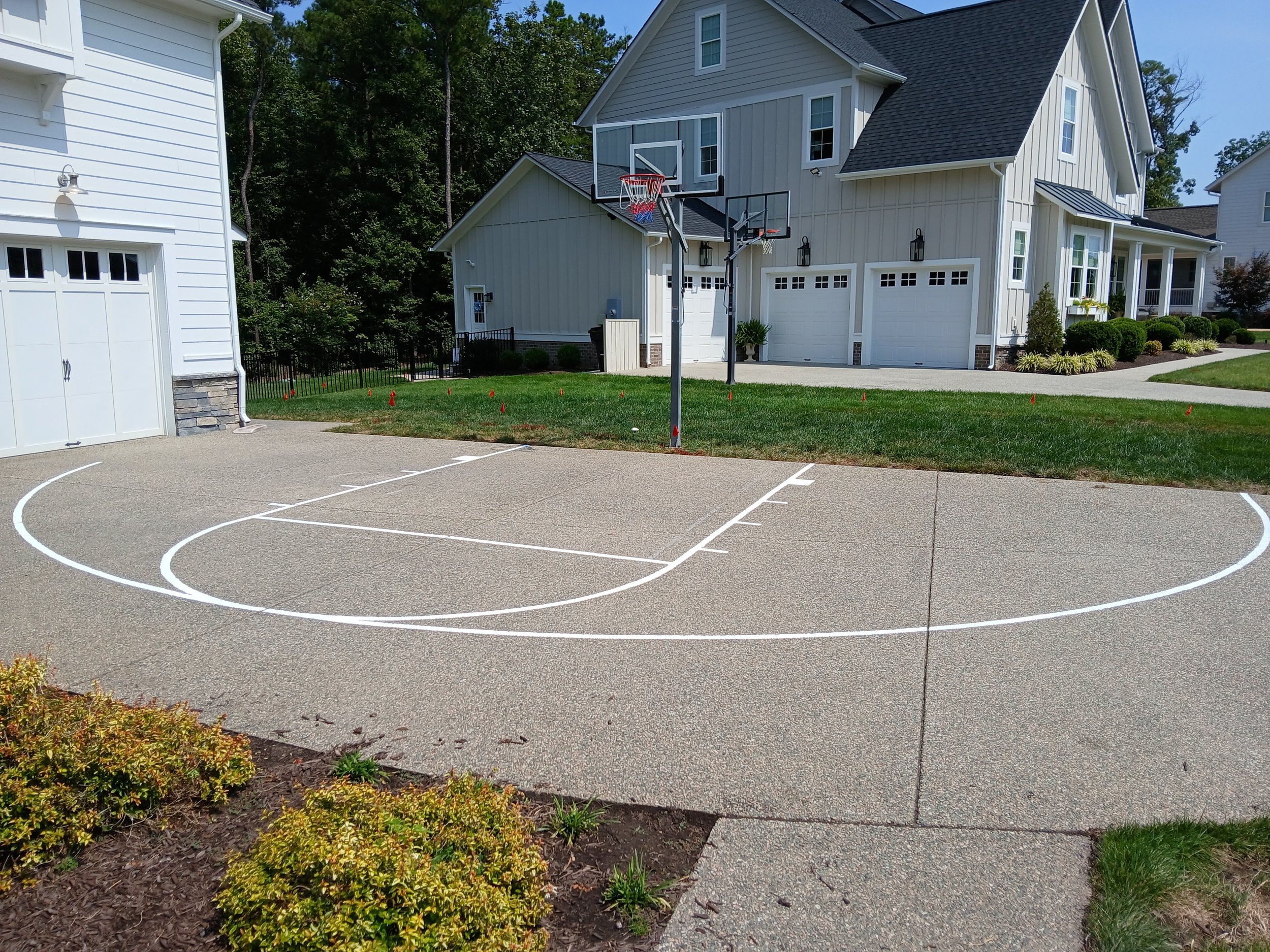 A driveway with a freshly painted basketball court outline in front of a modern house with a basketball hoop mounted on a pole.
